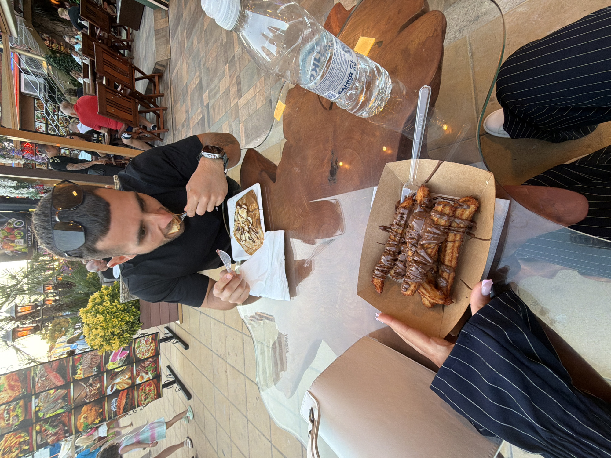In Nesebar, Bulgaria, two individuals are seated at a transparent, multi-level glass table outdoors. One person, a male with sunglasses on his head, is actively eating from a white plate with a fork, holding a plastic utensil. On the table, in front of him, is a plate with a crepelike dessert topped with nuts and drizzled chocolate. To the left of the plate, a white napkin is present. Next to the crepelike dessert, a partially empty plastic water bottle is positioned. In the foreground, a portion of another person's arm and hand, with painted nails, is visible, holding a brown cardboard container filled with churros drizzled with chocolate. A beige purse is also on the table near the churros. The background features a large, colorful menu board displaying various food items, a potted plant with yellow flowers, and a glimpse of other diners and restaurant seating, suggesting a dining establishment. The overall setting appears to be a warm, outdoor environment, possibly a restaurant terrace.