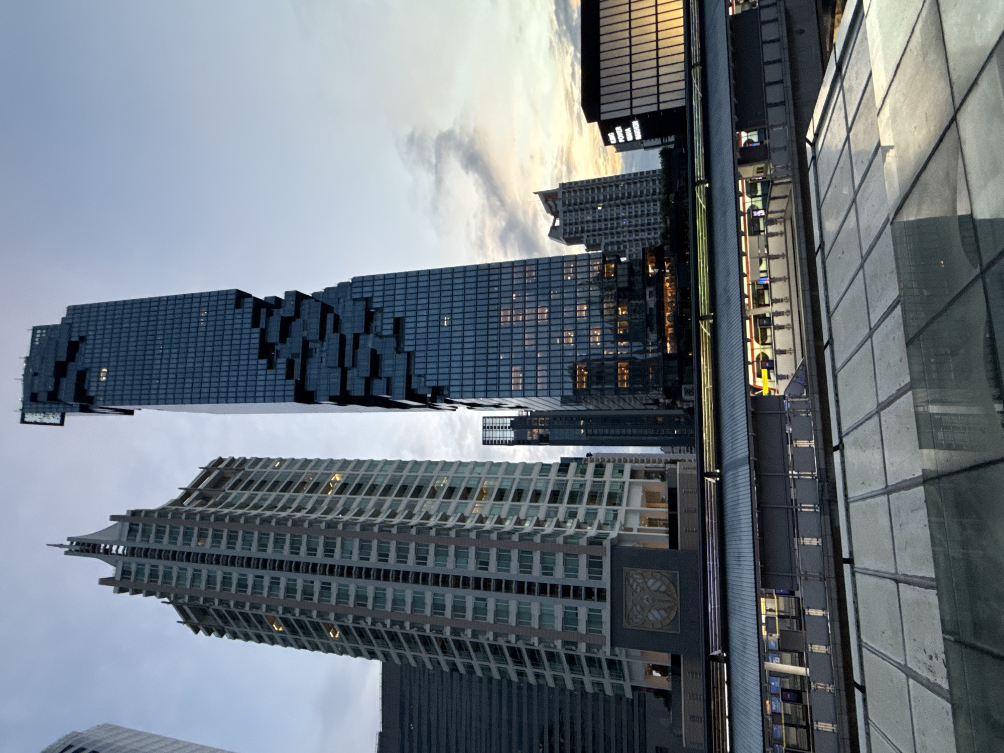 The image captures a twilight view of the King Power MahaNakhon building in Bangkok, Thailand. The distinctive pixelated facade of the skyscraper dominates the right side of the frame. To its left stands a taller, more conventionally designed skyscraper with a pointed spire, its balconies and windows illuminated. In the foreground, a modern elevated train station platform is visible, with trains docked and lights on. The sky is a blend of dusky blues and oranges, with scattered clouds indicating the transition from day to night. Reflective surfaces in the foreground, possibly glass or polished metal, mirror the scene above. The overall impression is an urban landscape at dusk, with contemporary architecture and transportation infrastructure.