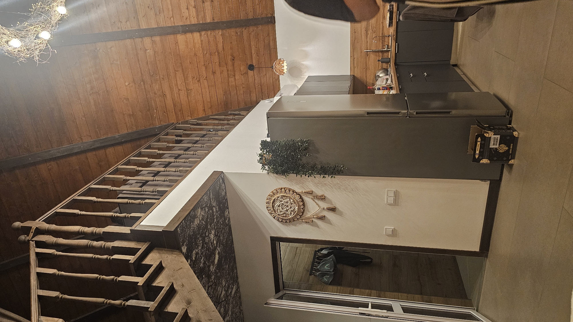 A slightly tilted, high-angle interior shot shows a staircase, a kitchen area, and decorative elements. The staircase, made of dark-stained wood, ascends from the lower left corner. A white wall runs adjacent to the staircase. A large decorative print, depicting a forest scene in shades of gray and black, is on the wall next to the staircase.
The kitchen area is on the right side of the image. It features gray cabinets and a refrigerator. A small decorative chest rests on the edge of the counter. Above the refrigerator, there is a small decorative plant. On the wall adjacent to the refrigerator, there is a woven wall hanging with tassels. Below it, there is a light switch and an outlet, and a window with a metal frame.
The ceiling and a portion of the wall above the staircase are covered in brown wood paneling. A decorative light fixture, resembling a bird's nest with lights, hangs from the ceiling.
The shot appears to be taken inside a residence. Without further information, it's impossible to confirm whether it is located in Ufa, Russia.