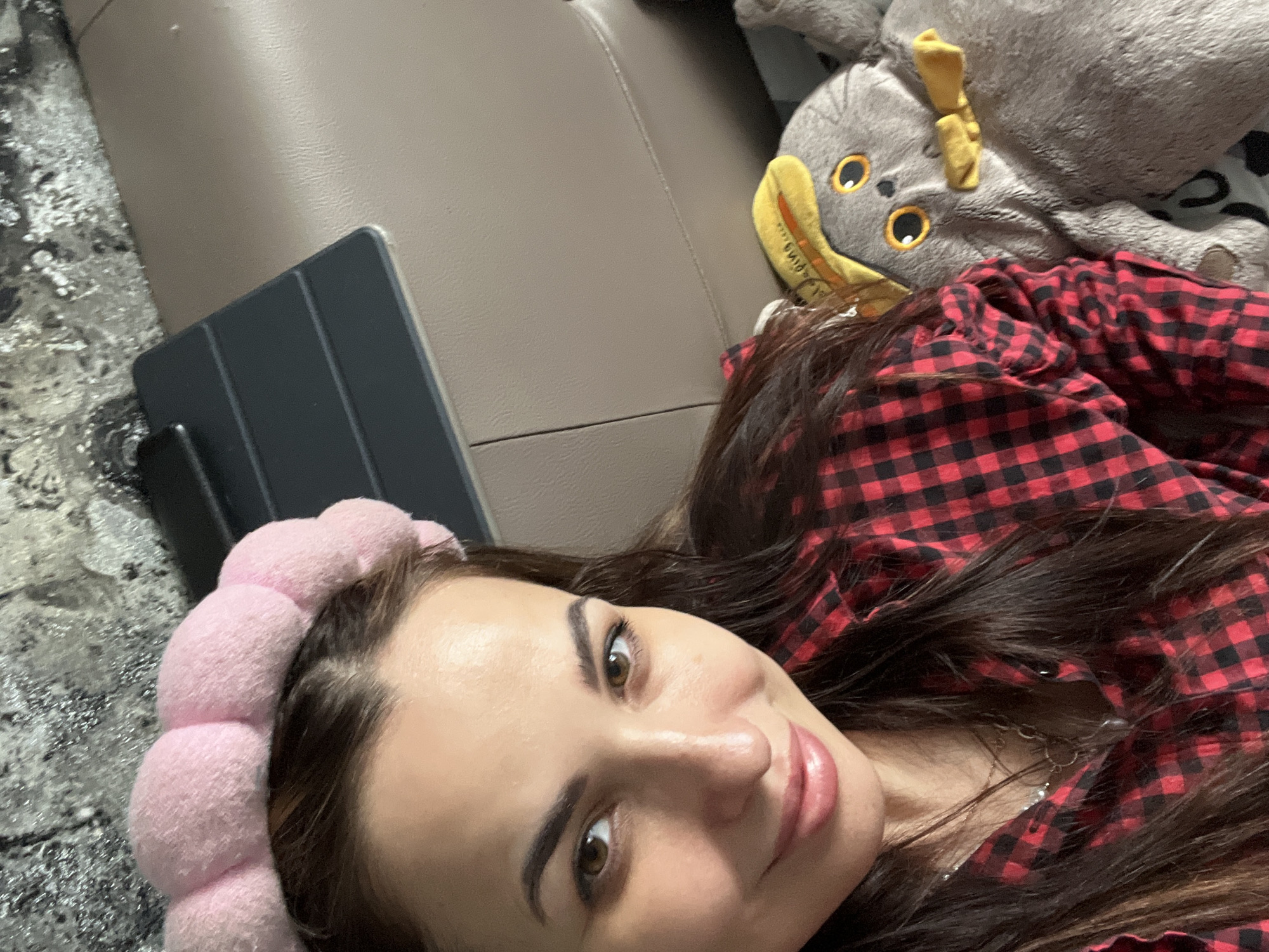 A person is reclining next to a gray stuffed animal with yellow features. The person is wearing a red and black checkered shirt and a pink puffy headband. A gray tablet lies on a gray-patterned surface next to a beige surface.