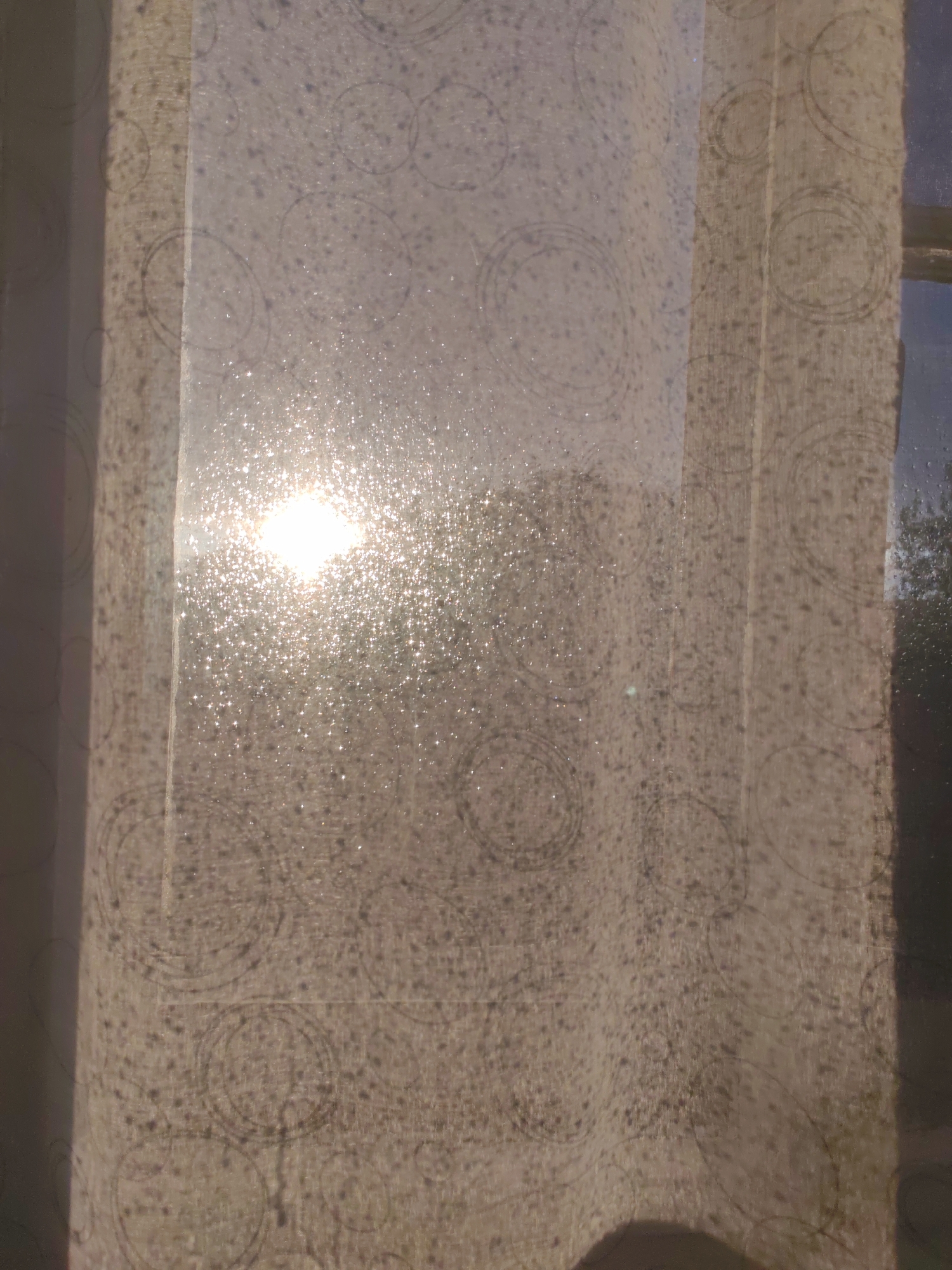 The image depicts a close-up view of a translucent, patterned curtain, likely made of light fabric, hanging in front of a window. The sun is shining brightly through the curtain, creating a dazzling effect with numerous small, sparkling highlights. This suggests it is daytime, and the sun is either rising or setting, given the warm, golden light.
The curtain has a subtle, repeating pattern of concentric circles and small dots, which are visible through the diffused sunlight. The fabric itself appears to be a light, neutral color, possibly beige or off-white. The sunlight is intense enough to cause some glare, making it difficult to discern details outside the window clearly. However, hints of trees and possibly a building can be seen through the fabric on the right side of the image, indicating an outdoor scene beyond the window.
The overall mood is serene and warm, evoking a sense of peacefulness associated with natural light. The focus is on the interplay of light and fabric, with the sparkling effect of the sunlight catching the texture of the material. No people or specific objects are prominently featured, the subject being the natural phenomenon of light filtering through a domestic setting. There is no visible text in the image. The location context of Сокол, Russia, provides a geographical setting for this scene of everyday life.