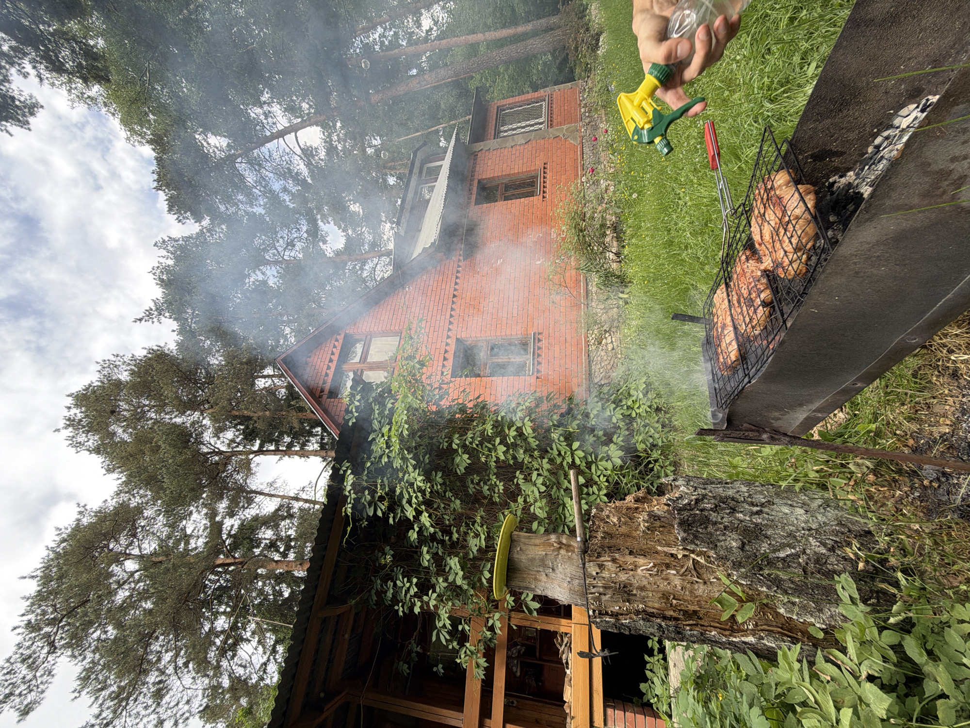 The image, taken outdoors, shows a man using a yellow and green spray bottle to extinguish flames from a metal grill where meat is being cooked. Smoke billows upwards. A two-story brick house is in the background, surrounded by trees and greenery. A wooden structure with a sloped roof is also visible to the side of the house. The location is likely Udelnaya, Russia, judging by the architectural style and surrounding environment.