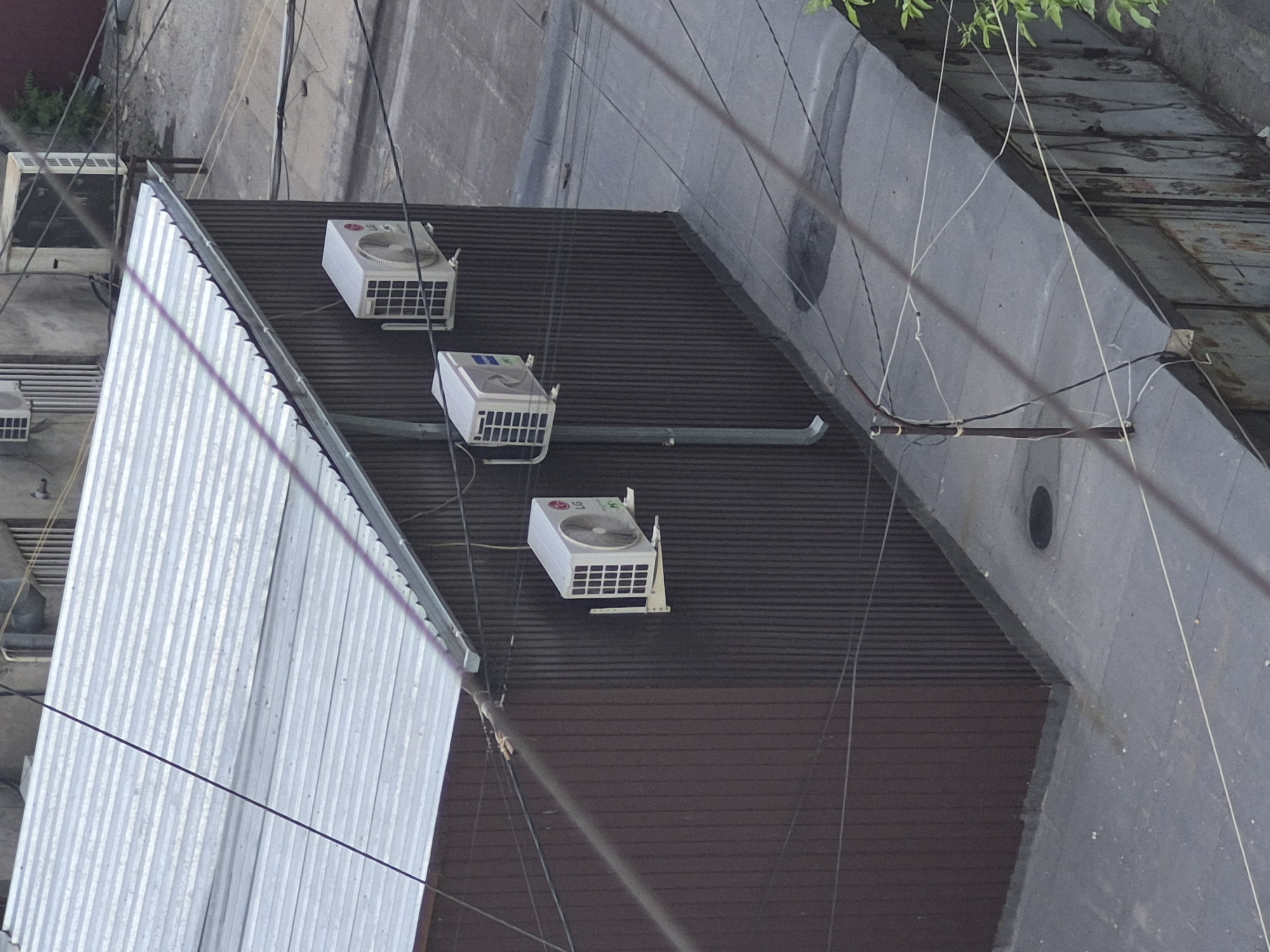 The image presents an angled, overhead view of a building's upper exterior or rooftop, characterized by industrial and utility elements. A significant portion of the scene is covered by a dark brown, ribbed surface, likely a roof or siding.
Mounted on this dark surface are three white, rectangular air conditioning condenser units. Two of these units clearly display the "LG" logo, featuring a fan on one side and a grilled vent on the front, supported by white brackets. A light grey drainage pipe or conduit extends horizontally along the ribbed surface, seemingly connected to one of the AC units.
To the left, a light-colored, corrugated metal structure, possibly a wall or secondary roof, rises vertically, featuring a jagged, pointed edge along its top, perhaps a bird deterrent. Another, older-looking air conditioning unit is partially visible near the top left, obscured by green foliage and a network of black and white electrical or communication wires that crisscross the entire frame.
In the background and to the right, a large, grey concrete wall or structure stands prominently, displaying texture and a dark circular opening. The lighting suggests it is daytime, but no specific weather conditions are indicated. There are no people visible, and no overt activities are taking place, portraying a static urban environment. The visible "LG" branding is the only legible text.