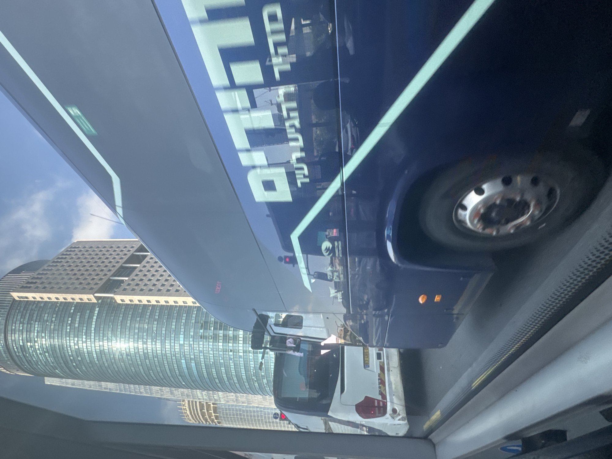 This image captures a view from inside a vehicle looking out at city traffic in Tel Aviv, Israel. The foreground features the interior of the vehicle with a reflective surface showing a partial view of the sky and a sliver of the environment outside. Prominently displayed on the side of a large, dark blue bus, is text in white Hebrew letters, part of which is legible as "מהיר להגיע רעננה" (Mahir LeHagia Ra'anana), translating to "Fast to reach Ra'anana". A lighter colored bus is visible in the middle ground, also in traffic. In the background, several modern, tall skyscrapers dominate the skyline, characteristic of a major city center. The sky is partly cloudy with a bright, sunny atmosphere. The traffic appears to be moving slowly, suggesting a possible traffic jam or congested road.