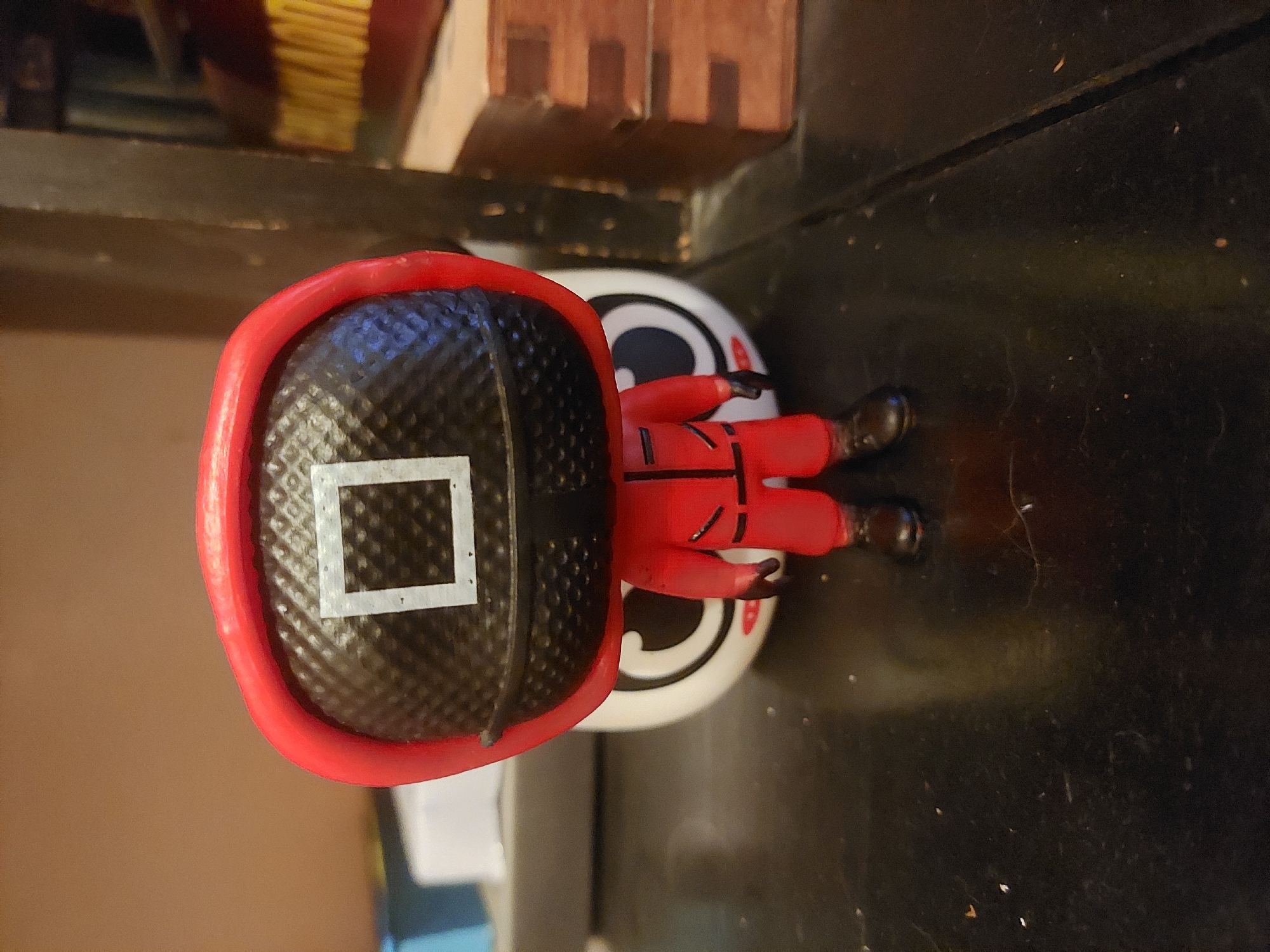 A close-up, slightly angled shot depicts a "Squid Game" themed figurine. The figure has a round white head with cartoonish eyes. Over the head is a red helmet with a square design on the front. The body is small and red. The background is a dark, grainy surface. The figurine is positioned on a dark surface with dust.
Tsarevo, Bulgaria is not evident from the image.