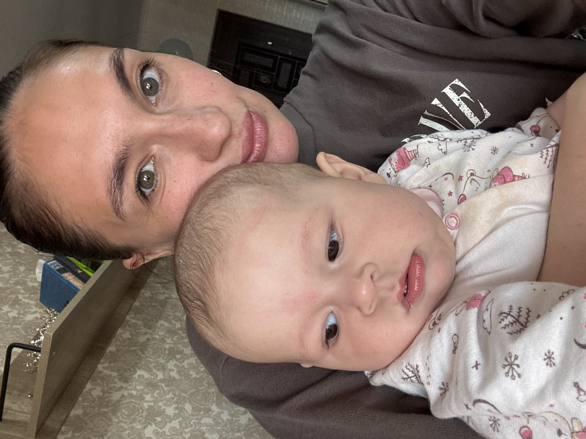 The image captures a close-up, intimate moment between a woman and a baby, likely a mother and her child, in an indoor setting in Sochi, Russia. The perspective suggests a selfie, with both individuals lying down or reclining.
The woman, positioned in the upper left of the frame, looks directly at the viewer with soft, light brown or hazel eyes. Her dark hair is pulled back, revealing clear skin and subtle, natural features. She wears a dark brown or grey long-sleeved top, which has a distinct white graphic logo on the chest; the logo appears to be a stylized letter, possibly an 'F' or 'E', with radiating lines.
Cradled against her, the baby occupies the center and right of the frame. The baby has fair skin, prominent rosy cheeks, and light brown hair. Its dark eyes are open and directed slightly to the right, and its mouth is gently open. The baby is dressed in a white long-sleeved onesie decorated with cheerful patterns in pink, brown, and black, including snowflakes, bears, hearts, and crosses.
The background details suggest a home environment. A dark, possibly wooden, furniture piece is visible behind the woman. In the lower left, a light-colored wooden shelf holds a blue box and another item, along with a black metal stand. The floor or carpet underneath has a subtle patterned design. The overall scene conveys a sense of peaceful connection and affection between the mother and child.