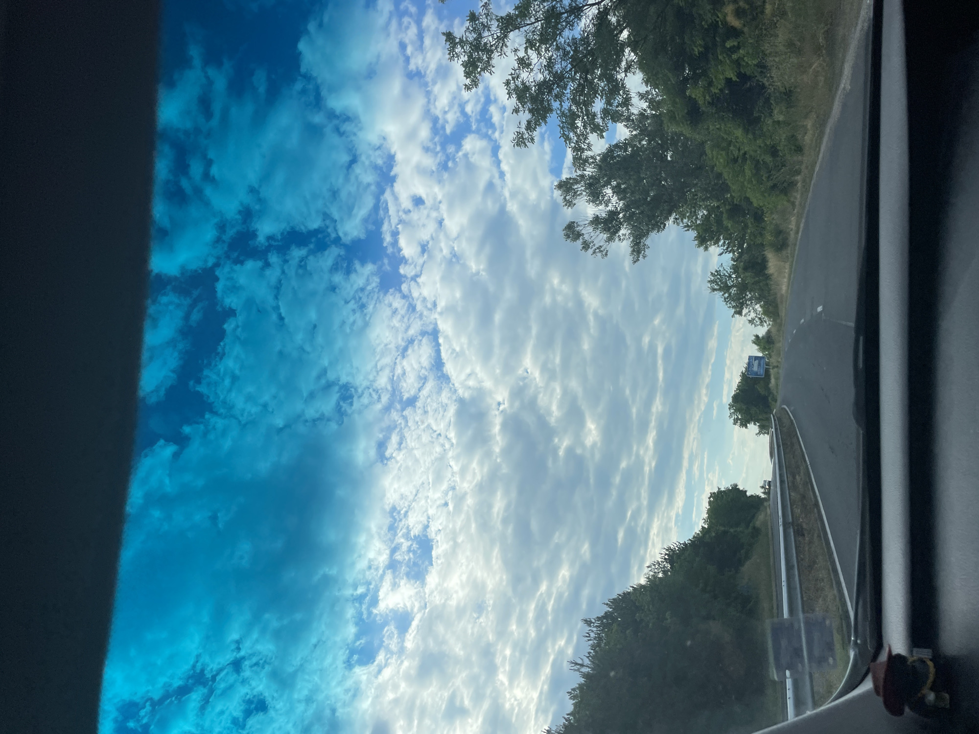 The image captures a view from inside a vehicle traveling on a road that appears to be a highway or a major route. The road is paved and marked with white lines. On either side of the road are lush green trees and vegetation, suggesting a rural or semi-rural environment. The sky is a prominent feature, filled with dramatic white and gray clouds against a bright blue sky, indicating it might be a partly cloudy day with good visibility. The foreground shows the dashboard of the vehicle, with a small decorative item that resembles a cowboy figurine with a hat. In the distance, there are signs and a few vehicles, suggesting the presence of other traffic. There is a guardrail visible along the side of the road. The overall impression is of a journey through a scenic landscape under a dynamic sky. The user prompt mentions the city of Balabanovo, Bulgaria, but there are no specific visual cues within the image to confirm this location.