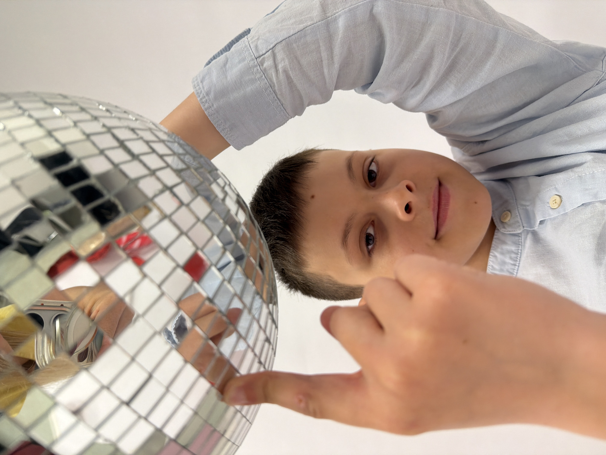 A young boy in a light blue button-down shirt is angled towards the camera, holding a disco ball. His right index finger is pointing towards the disco ball. The background is a plain white. There is no visible information in the image to suggest the location of the subject or image capture.
