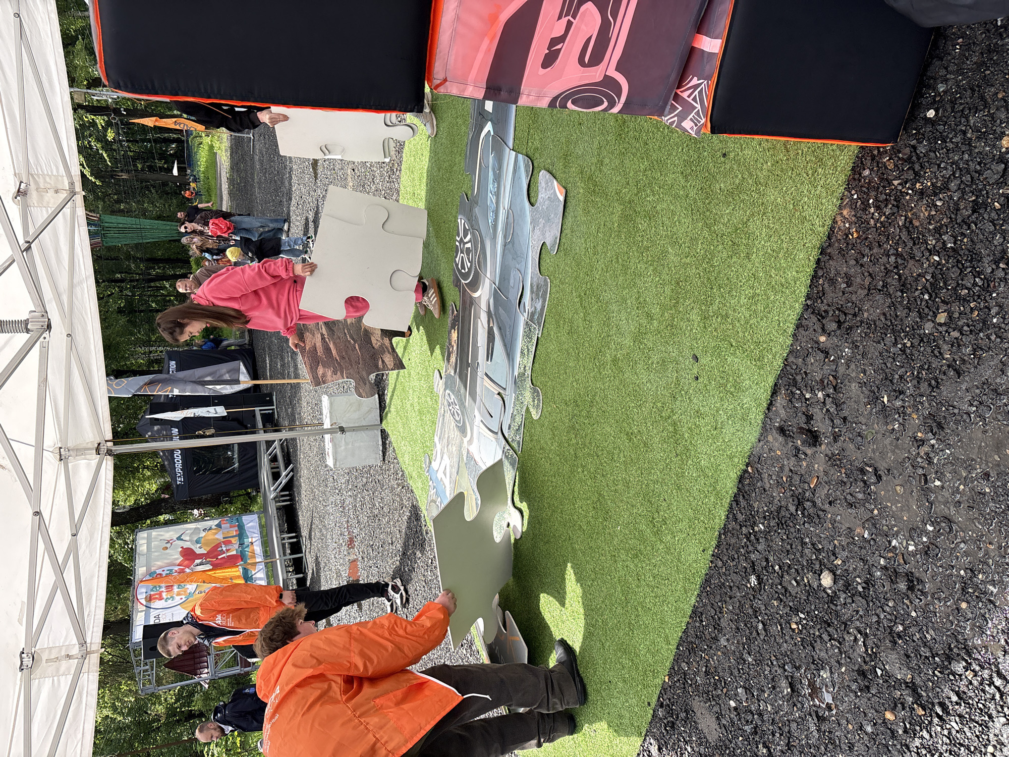 A group of people are engaged in assembling a large jigsaw puzzle on a green artificial turf. The puzzle image appears to be a vehicle. One person is wearing a pink sweatshirt and another an orange jacket. In the background, a crowd of people is visible, along with event-related banners and displays. The location is outdoors and appears to be in Izhevsk, Russia. The ground adjacent to the artificial turf is asphalt.