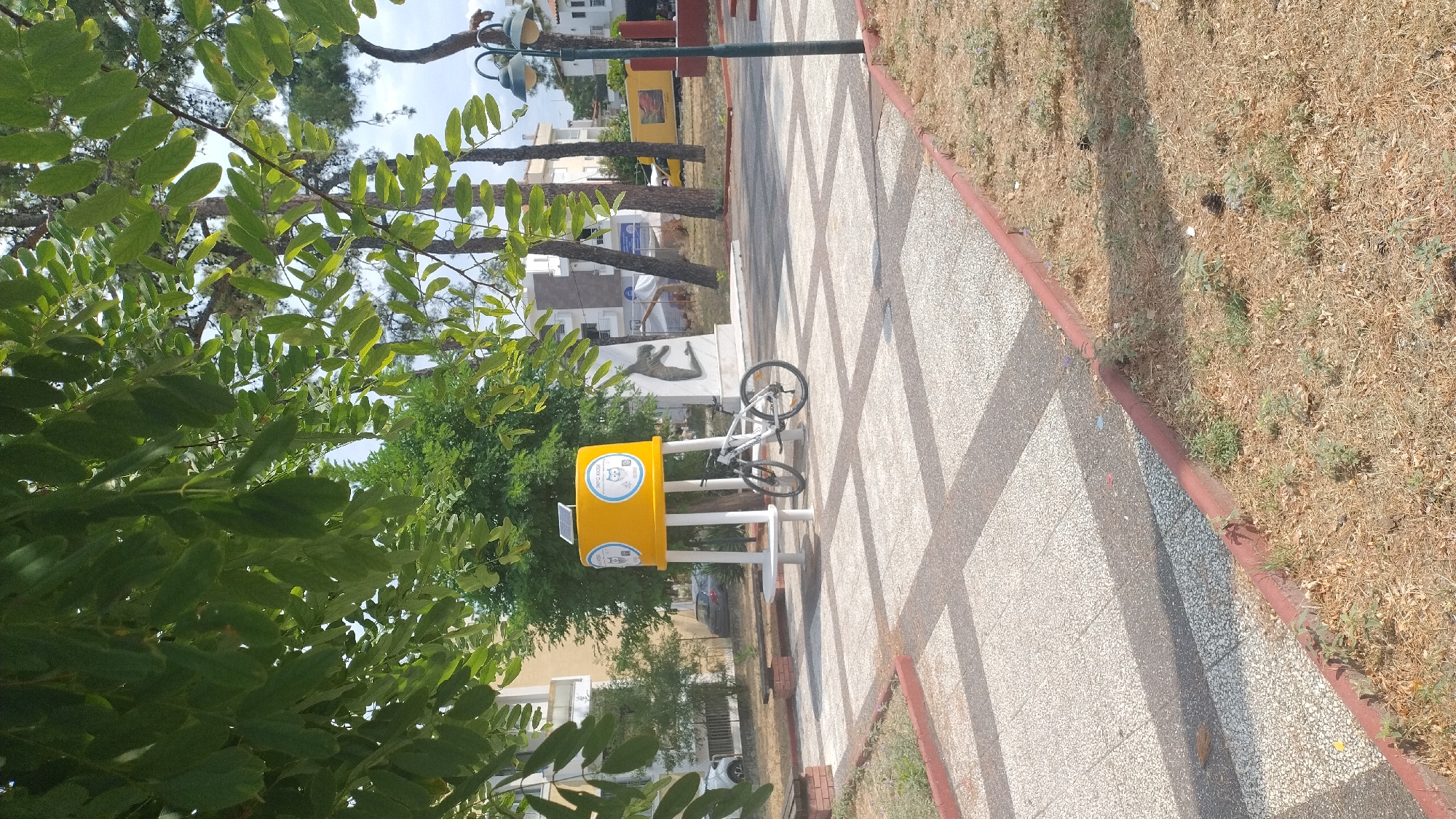 An outdoor scene in Neoi Epivates, Greece shows a paved plaza area with a modern-looking yellow cylindrical structure supported by white poles. The structure has a solar panel on top and two circular emblems with cat graphics and Greek text. A white bicycle is parked next to the structure. The plaza is bordered by a red curb and dry grass. Trees with green leaves overhang the scene, casting shadows on the ground. In the background, there are buildings, a lamp post, and a large stone sculpture of a human figure. The sky is partly cloudy.
