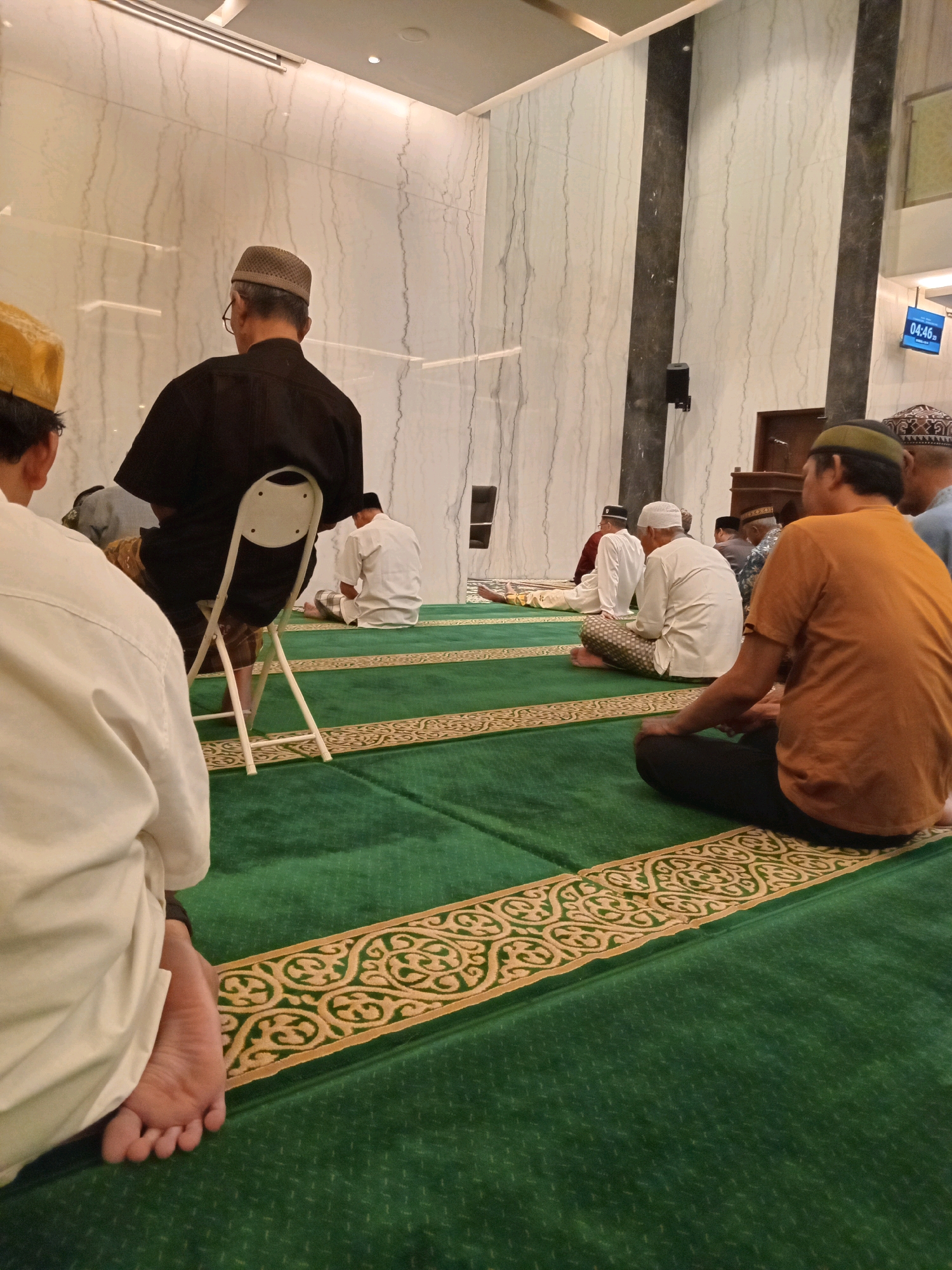 The image shows a group of men sitting on green prayer rugs inside a mosque. The mosque has white marble walls with dark vertical accents. There is a digital clock on the wall displaying "04:46". Most of the men are wearing traditional Islamic attire, including caps and long robes. Some are seated cross-legged, while others are on chairs or in prayer positions. The rugs have a decorative border. The scene appears to depict a prayer gathering. The location is identified as the city of Randuagung, Indonesia.