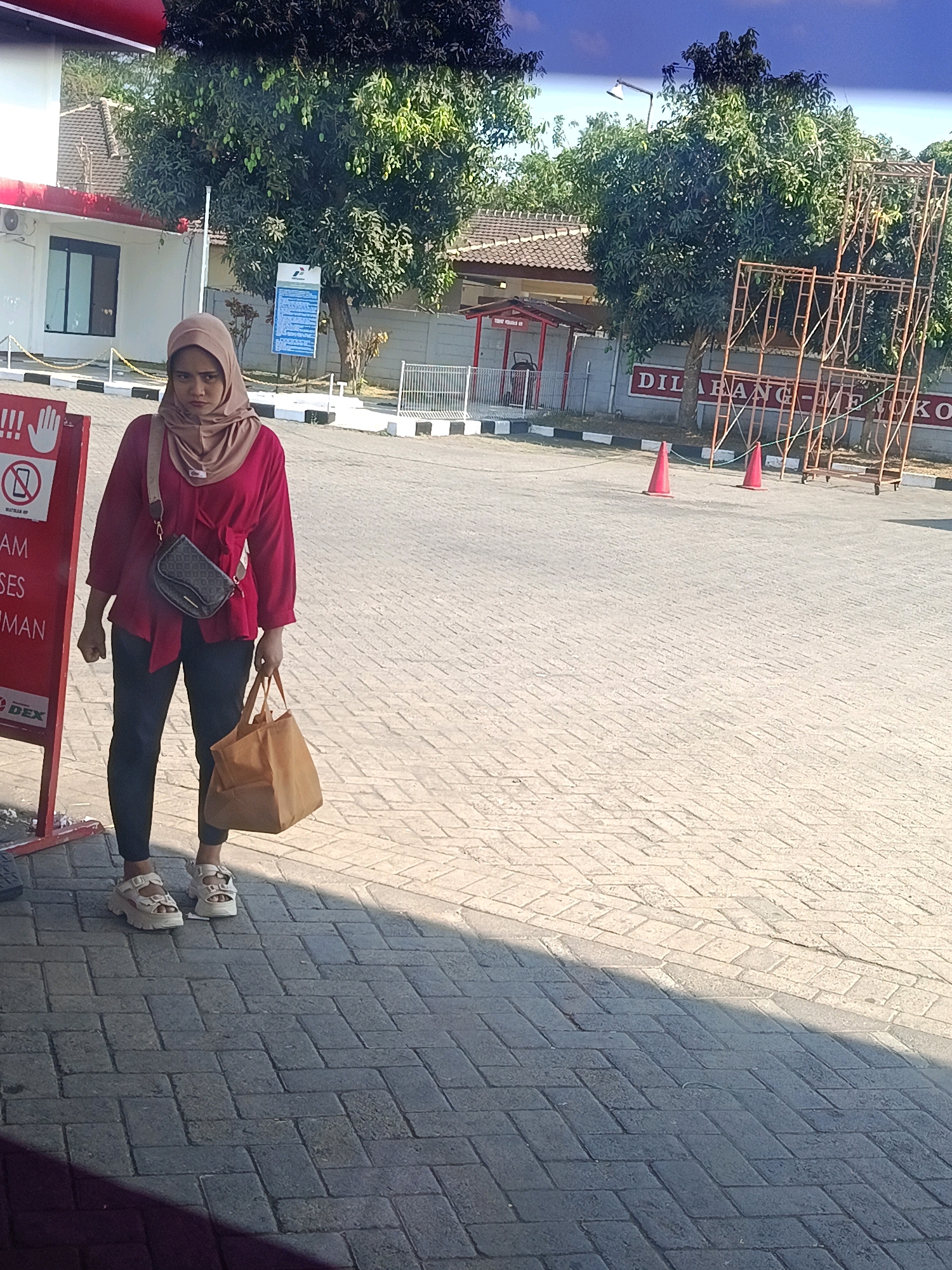 A woman stands on a paved area in front of a building. She is wearing a light brown hijab, a fuchsia shirt, and dark pants. She carries a small dark bag across her body and a larger light brown tote bag in her right hand. In the background, there is a white building with a red awning and a sign that reads "DILARANG MEROKO". There are also trees, a scaffolding structure, and two orange traffic cones on the ground. The scene appears to be outdoors during the day. The building with the sign is located in Gedungjajang, Indonesia.