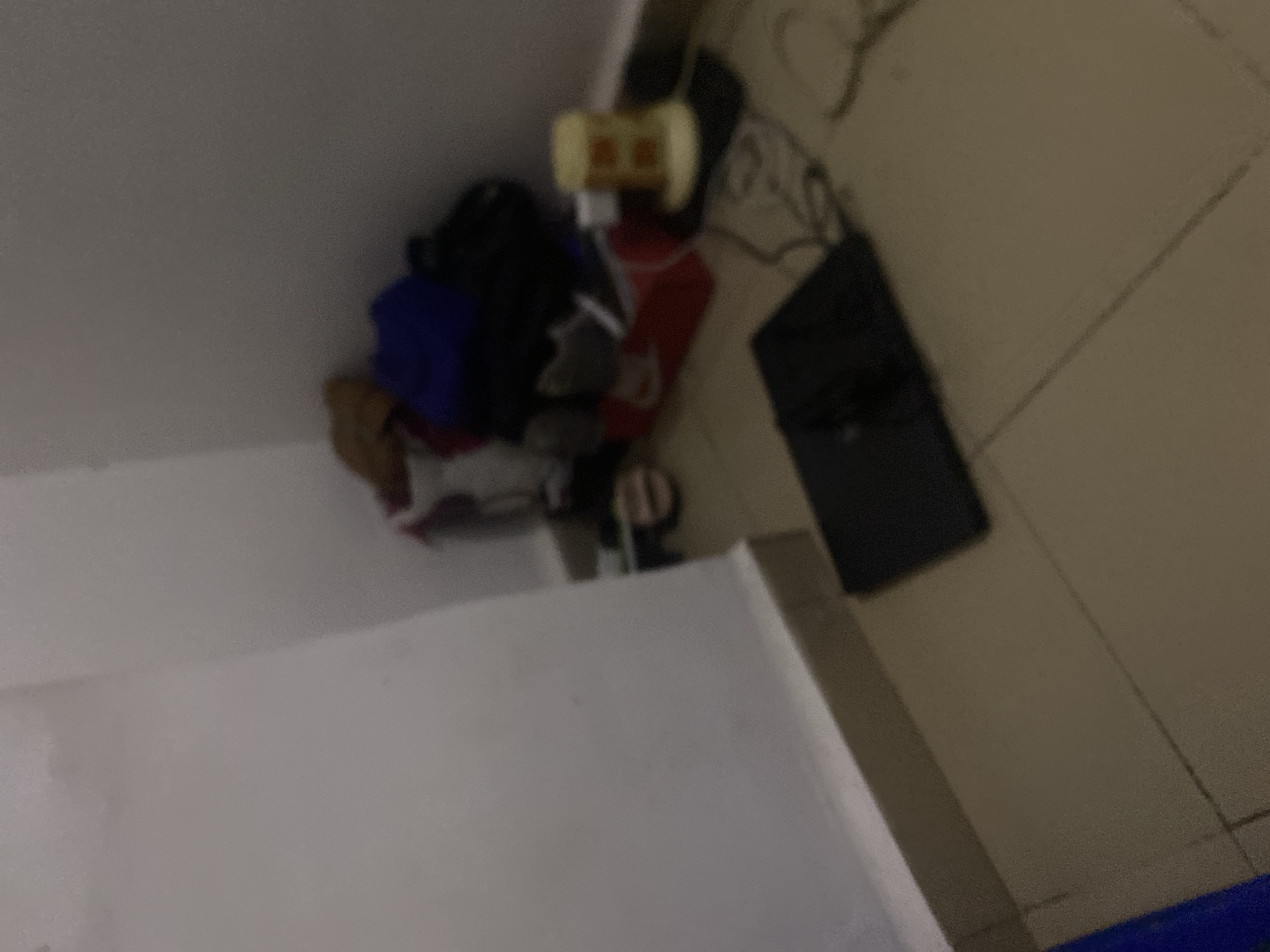 The image shows a dimly lit corner of a room, likely an apartment, in the city of Howayi, Nigeria. The floor is tiled in a neutral color, and the walls are painted white, with a beige baseboard along the bottom. In the corner, a pile of assorted items is gathered, including what appear to be bags, clothing, and possibly shoes. Among these items, a red box and a yellow cylindrical object are visible. In the foreground, a black electronic device, possibly a laptop or tablet, rests on the floor with cables connected to it. The overall impression is one of a cluttered living space.