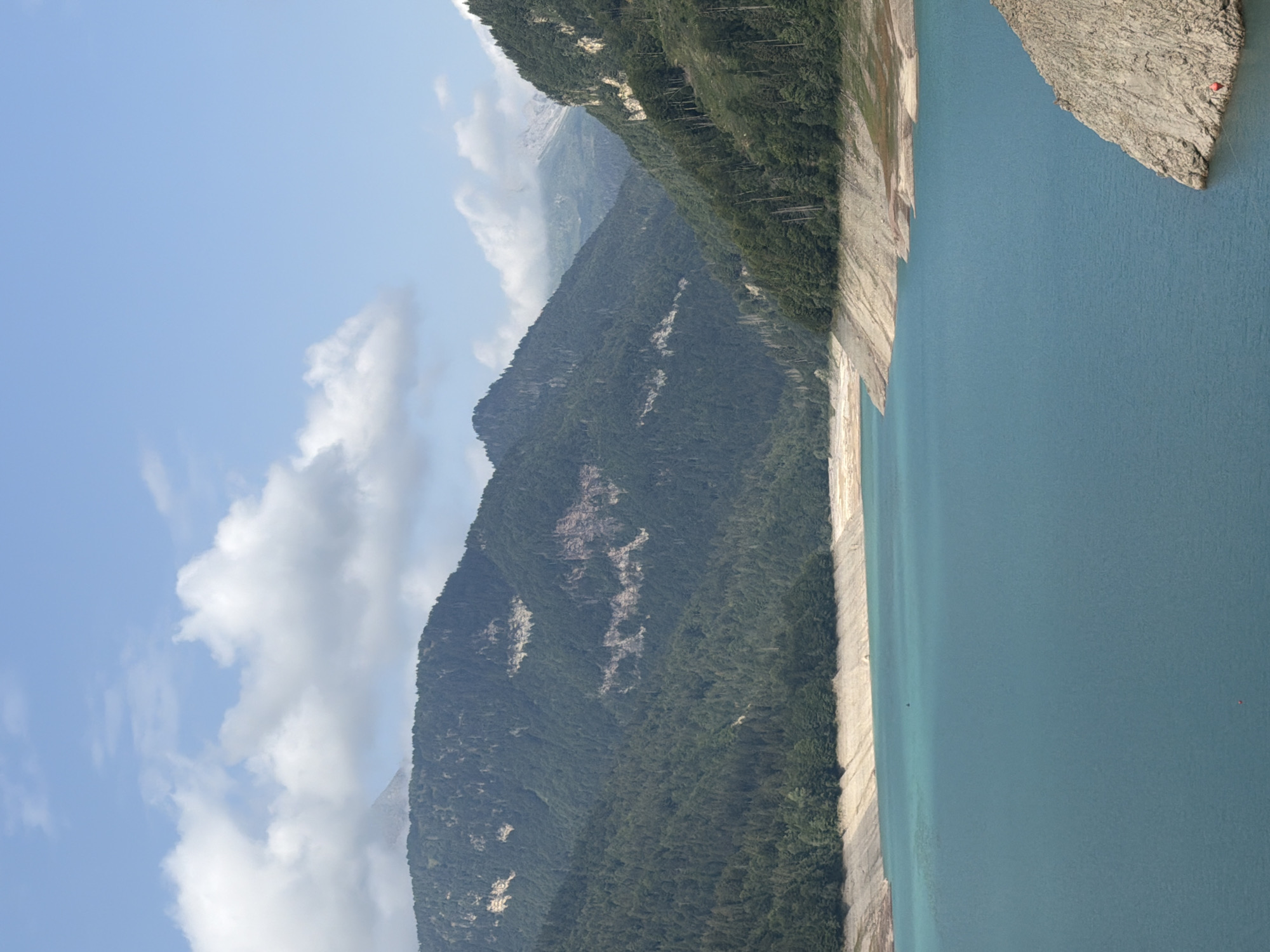 This is a vertical, high-angle shot of a vast, turquoise lake in a mountainous region. The lake occupies the lower two-thirds of the frame, with its surface calm and reflective. The shores of the lake are characterized by steep, rocky slopes with exposed, dry earth and scattered vegetation. In the background, towering mountains covered in dense green forests rise towards a partly cloudy blue sky. The overall impression is one of natural grandeur and a remote, pristine landscape. The scene is uninhabited by people. The location is the Ampezzo/Dimpeç area of Italy.