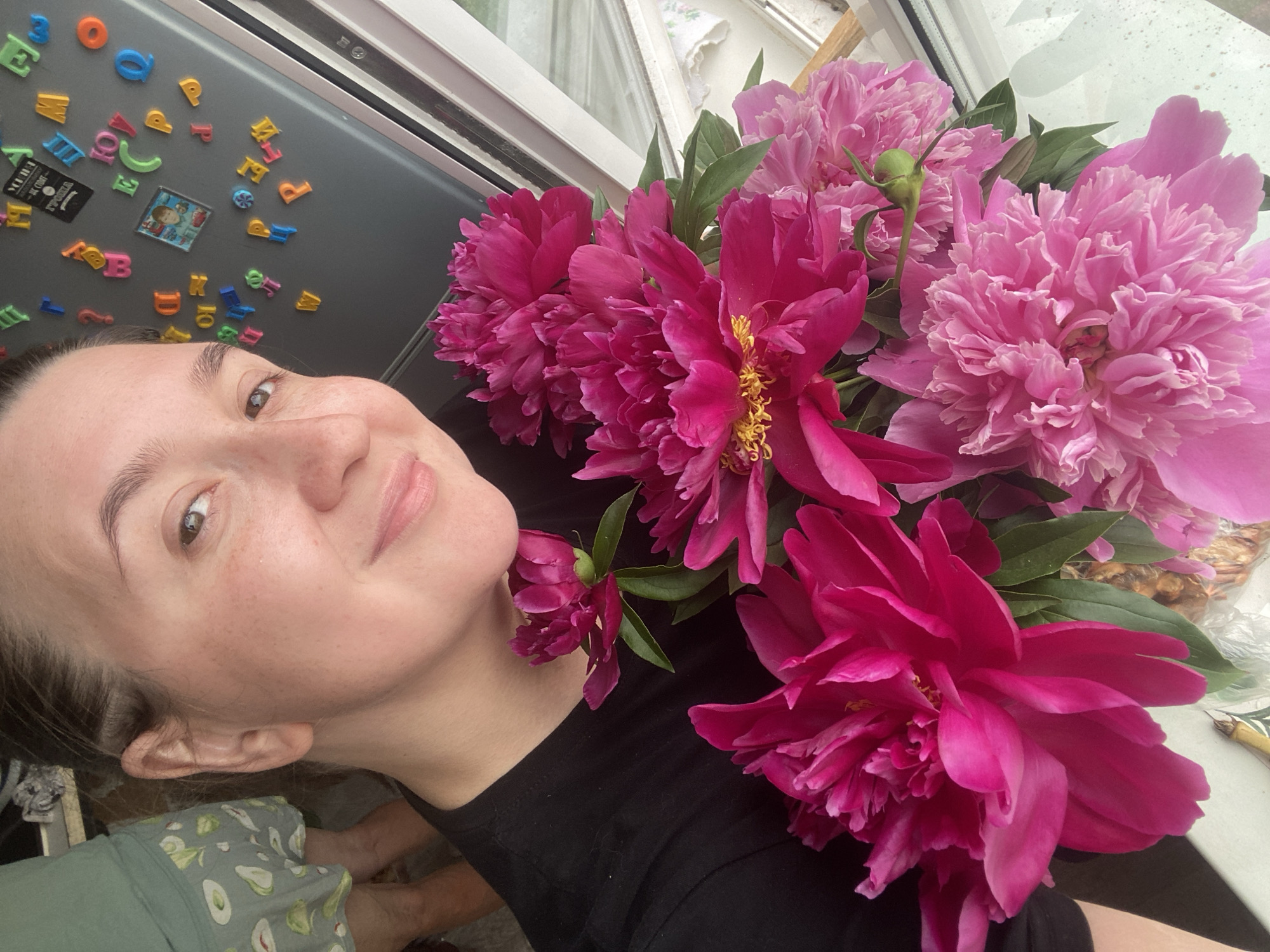 A woman is holding a bouquet of pink peonies. She is wearing a black t-shirt. Behind her, there is a refrigerator with magnetic letters on it, suggesting the scene might be in a kitchen or dining area. The presence of a window suggests it could be daytime. The overall setting appears to be domestic.