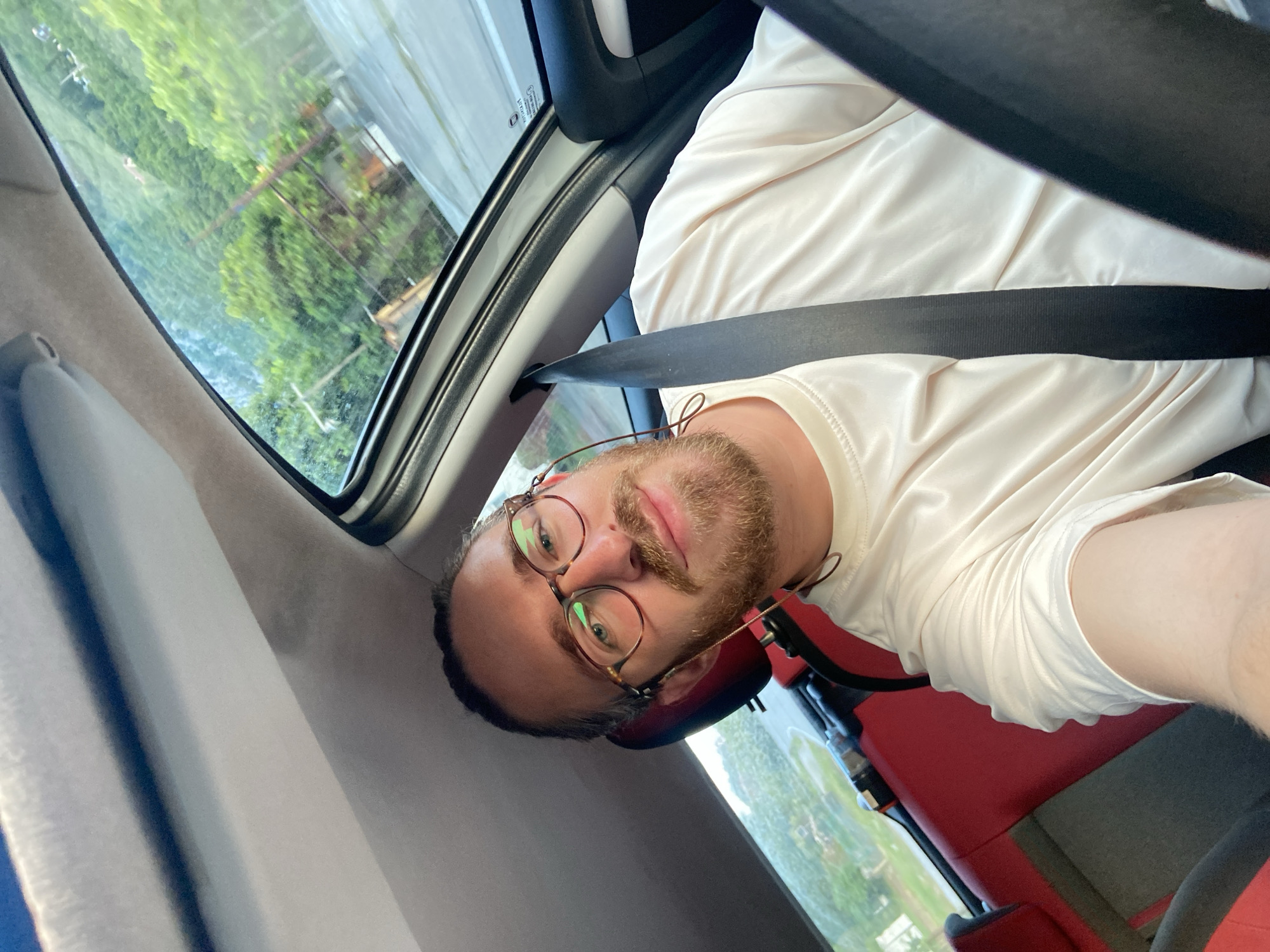 A man with fair skin, a reddish-brown beard, and round glasses is in the driver's seat of a car. He is wearing a white shirt and has a seatbelt across his chest. The car's interior is gray and red. Through the car window, green trees and glimpses of buildings can be seen. The image appears to be taken from a low angle, looking up and slightly angled. The location is Đurmani, Montenegro.