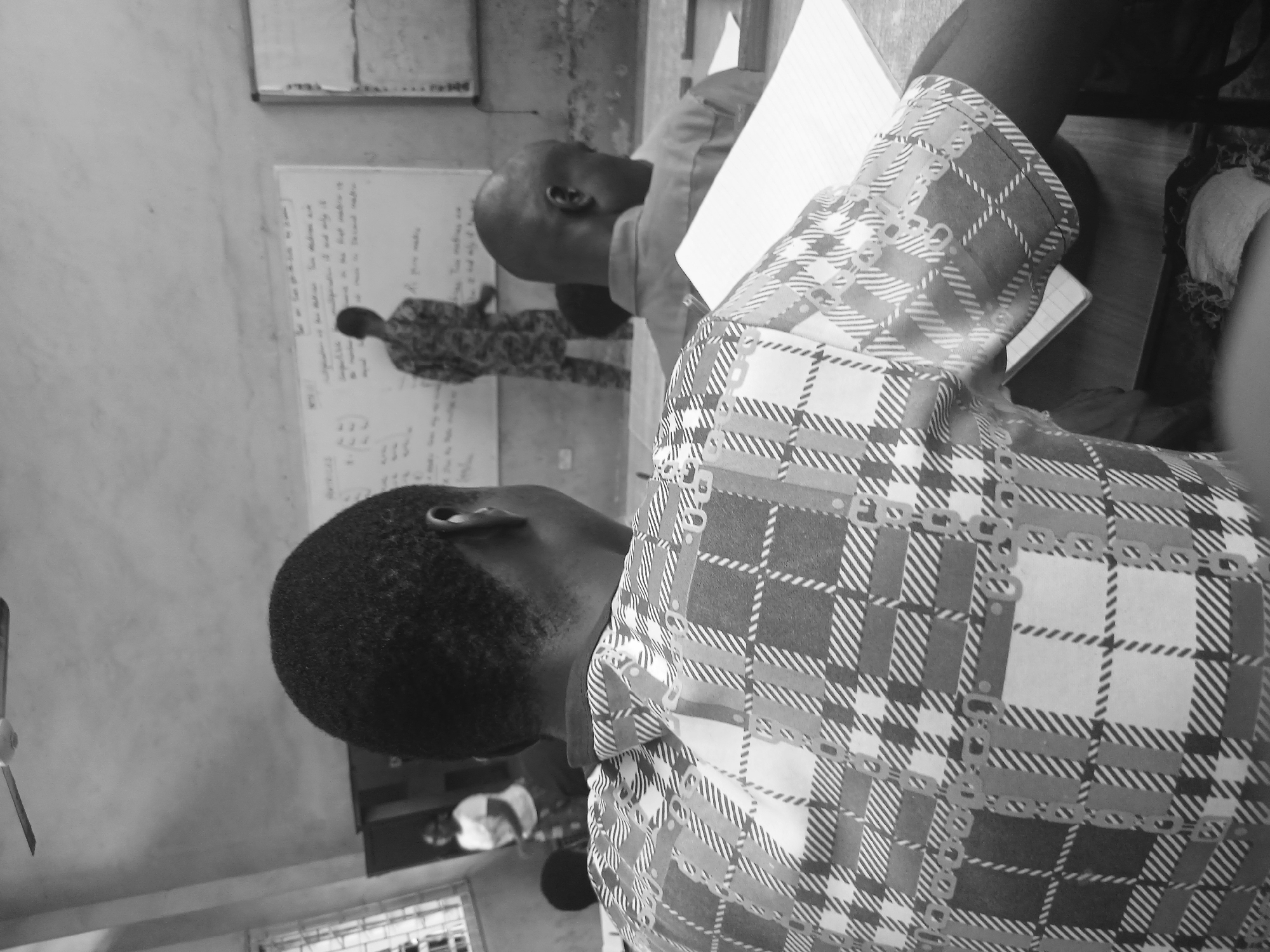 This is a black and white, medium shot, eye-level, indoor photograph taken in a classroom in Lagos, Nigeria. In the foreground, a student wearing a plaid shirt is seated at a desk, facing away from the camera. The student's head is visible from the back, and their right arm rests on the desk, with a notebook in front of them. To the right and slightly behind the foreground student, another student, with a bald head, is also seated at a desk and facing away from the camera. In the background, a person in a camouflage uniform is standing by a whiteboard that has writing on it. To the left of the person in uniform, a woman in a white t-shirt and patterned skirt is standing. The classroom appears to have a fan mounted on the ceiling and barred windows on the left side of the frame. The walls are bare and textured, and the overall lighting suggests an overcast day or an indoor artificial light source.