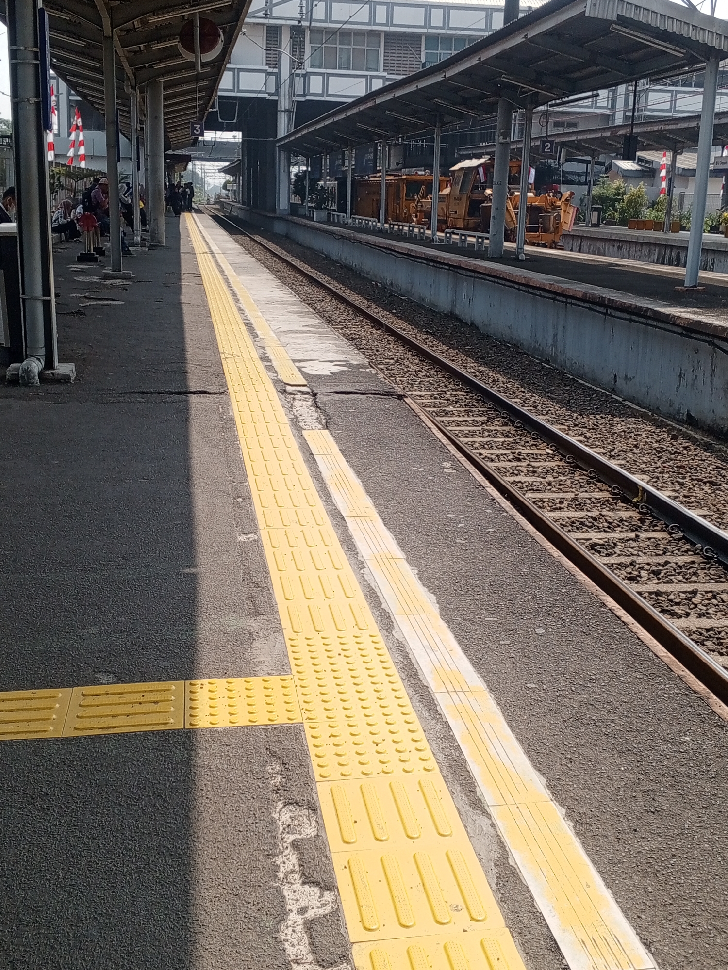 This image depicts a train station platform in Depok, Indonesia. The platform is paved with asphalt and features prominent yellow tactile paving for visually impaired individuals, running along the edge nearest the tracks. To the left of the tactile paving, the asphalt is shaded, creating a stark contrast with the sunlit area. In the background, a covered waiting area with pillars is visible, where several people are seated. Further down the platform, a train track runs parallel to the waiting area, with a concrete barrier separating the platform from the ballast. Behind the track, a grey concrete retaining wall supports the platform's edge. In the distance, a building with white framed windows and a metallic roof structure suggests the presence of station facilities. Parked on a separate track beyond the main line are two yellow industrial vehicles, possibly maintenance equipment, with one bearing a national flag. The sky is bright, indicating daylight.