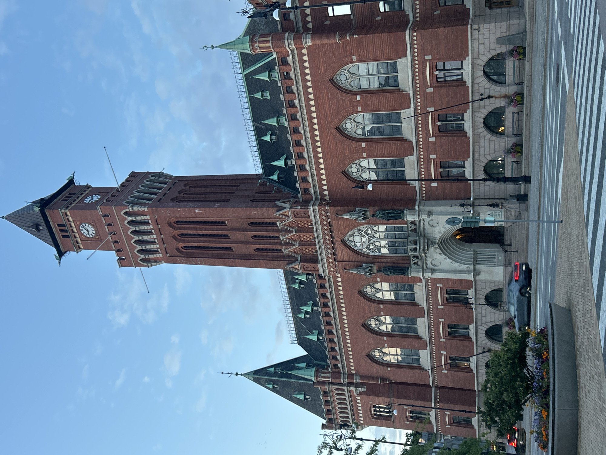 The image shows a frontal, low-angle view of the Helsingborg City Hall in Helsingborg, Sweden, during the day. The building is constructed primarily of red brick with green accents on the roofs and spires. A prominent clock tower rises from the center of the building, featuring clock faces on multiple sides. Arched windows with decorative stonework line the facade. In the foreground, a street with a pedestrian crossing is visible, with a dark-colored car driving away from the viewer. A circular planter with colorful flowers is situated on the left side of the street, and a tree grows within it. Streetlights are present on the sidewalk. The sky is partly cloudy, with a mix of blue sky and soft white clouds. The scene appears to be captured in natural daylight, with no artificial lighting evident on the building.