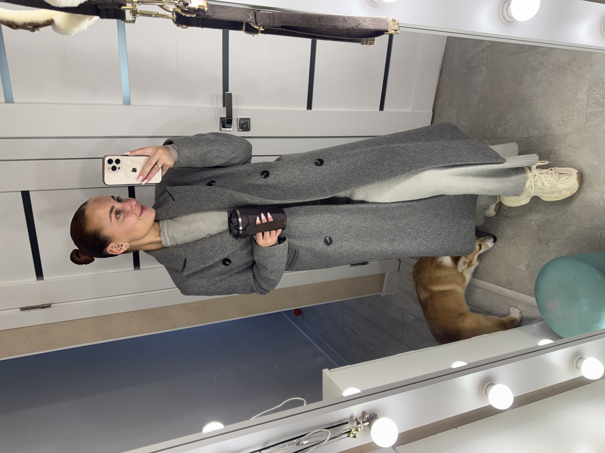 The image, currently rotated 90 degrees clockwise, depicts a woman taking a mirror selfie in what appears to be a modern indoor setting, likely a dressing room or bedroom, in Chaikovsky, Russia.
The woman, with her hair neatly pulled back, stands in front of a large vanity mirror illuminated by a strip of round, bright light bulbs. She is dressed in a long, grey wool overcoat, a light grey top, and light grey sweatpants, complemented by chunky cream-colored sneakers. In her right hand, she holds a light-colored iPhone, aimed to capture her reflection, and in her left hand, she holds a dark-colored camera or camcorder. She looks towards her phone with a neutral expression.
On the floor to her right, a fawn-colored Corgi dog is visible, lying down. A light blue/green cylindrical stool or ottoman is also partially seen next to the dog. The floor is covered with light grey tiles. The background reflected in the mirror shows white walls, a white door with dark handles, and what appears to be a dark leather belt with brass hardware hanging above. The smartphone shows a partial Apple logo and a MagSafe ring on its back. The setting is clean, well-lit, and contemporary.