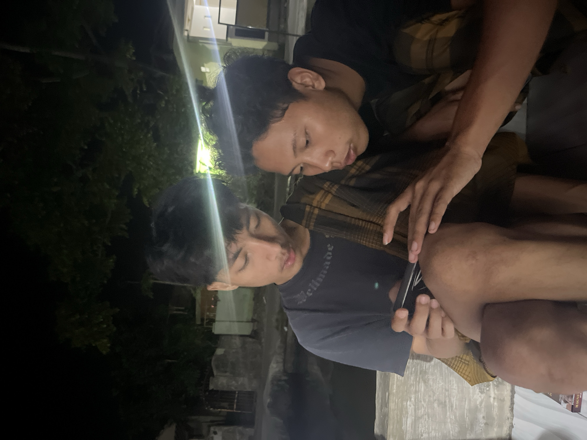 Two young men are sitting close together, one on the left and one on the right, in what appears to be an outdoor setting at night. The man on the left is wearing a dark t-shirt and a checkered cloth is draped over his shoulders and lap. He is holding a dark-colored object, possibly a phone, in his hands and looking down at it. The man on the right is wearing a black t-shirt and is leaning in, looking at the object in the other man's hands. The background shows trees and some structures, suggesting an urban or semi-urban environment. There are lights visible in the background, creating some glare and light streaks across the image. The scene is dimly lit overall.