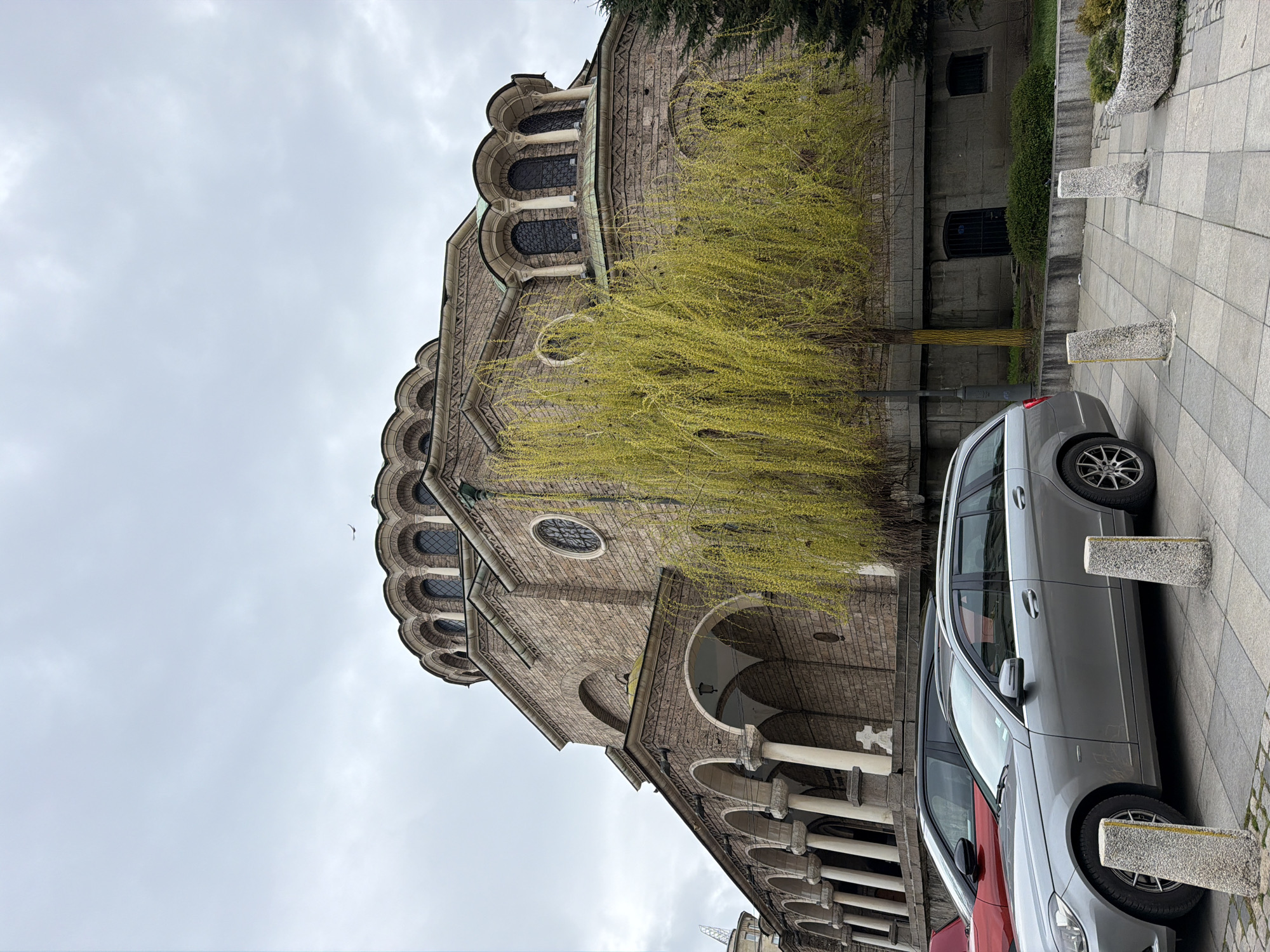 A low-angle shot of the St. Alexander Nevsky Cathedral in Sofia, Bulgaria, shows the structure on a cloudy day. A silver car is parked along the side of the cathedral next to the paved sidewalk with short, square bollards. A tree with bright yellow budding leaves partially obscures the stone facade of the cathedral. The architectural details, including arched windows, colonnades, and decorative stonework, are visible.