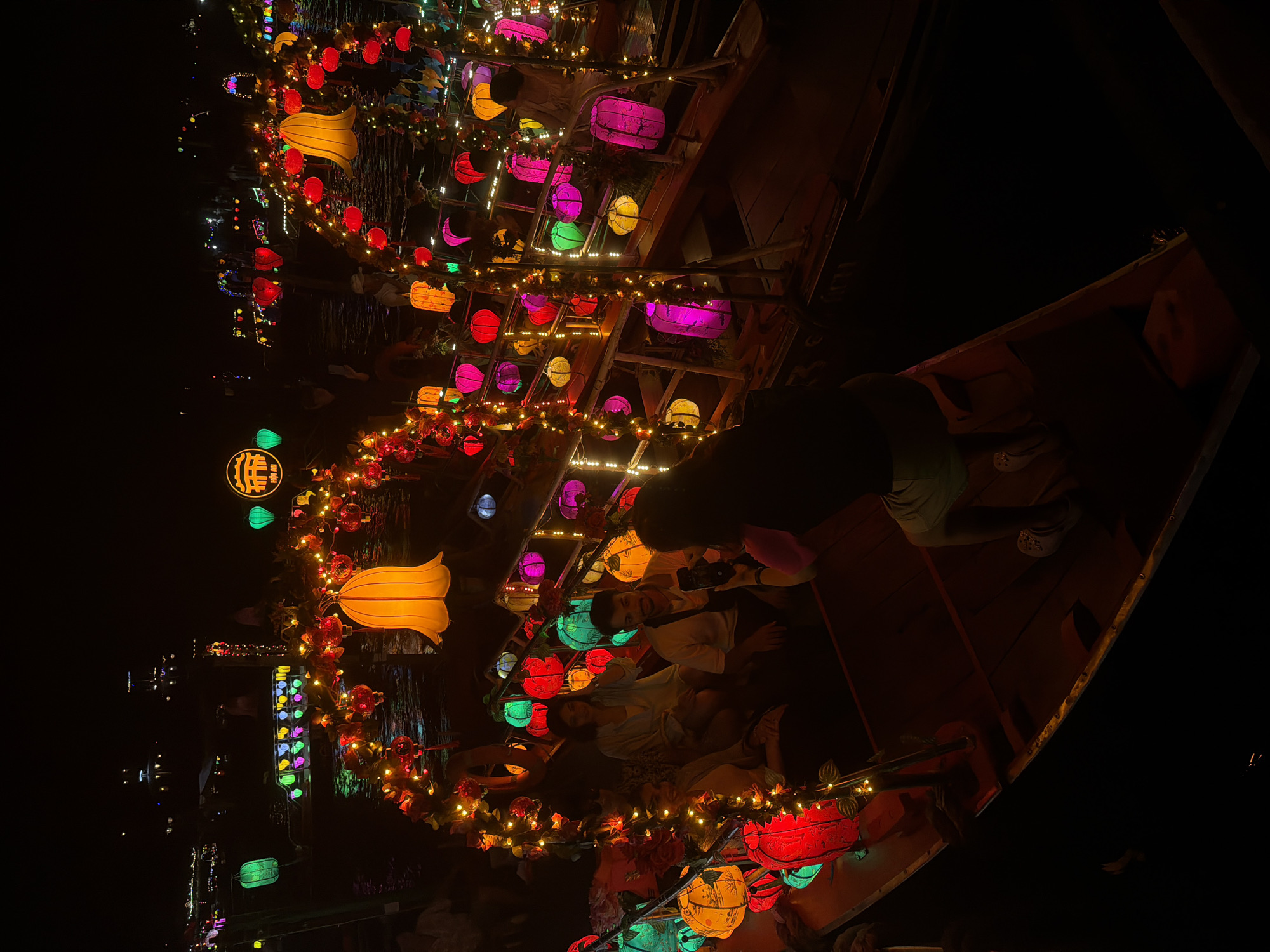 The image depicts a nighttime scene in Hoi An, Vietnam, likely during a festival or special event. A long wooden boat is filled with people who appear to be enjoying a leisurely ride. The boat and its surroundings are illuminated by a multitude of colorful lanterns, creating a festive and magical atmosphere. These lanterns, in various shapes and sizes, are strung along the sides of the boat and draped from overhead structures, casting a warm glow on the water and the faces of the people.
The people in the boat are seated and some are looking at the lanterns, while others appear to be conversing. One person in the foreground, wearing a light-colored shirt and green shorts, is leaning back and looking towards the rear of the boat. The boat itself is dark wood, with visible seating and a raised edge. The water reflects the vibrant lights, adding to the visual spectacle. In the background, more lanterns and lights can be seen, suggesting the event is spread across a body of water, possibly a river or canal. The overall mood is joyful and serene, a classic Hoi An nighttime experience. There is a circular lantern with what appears to be Chinese characters on it, hanging on the left side of the image.