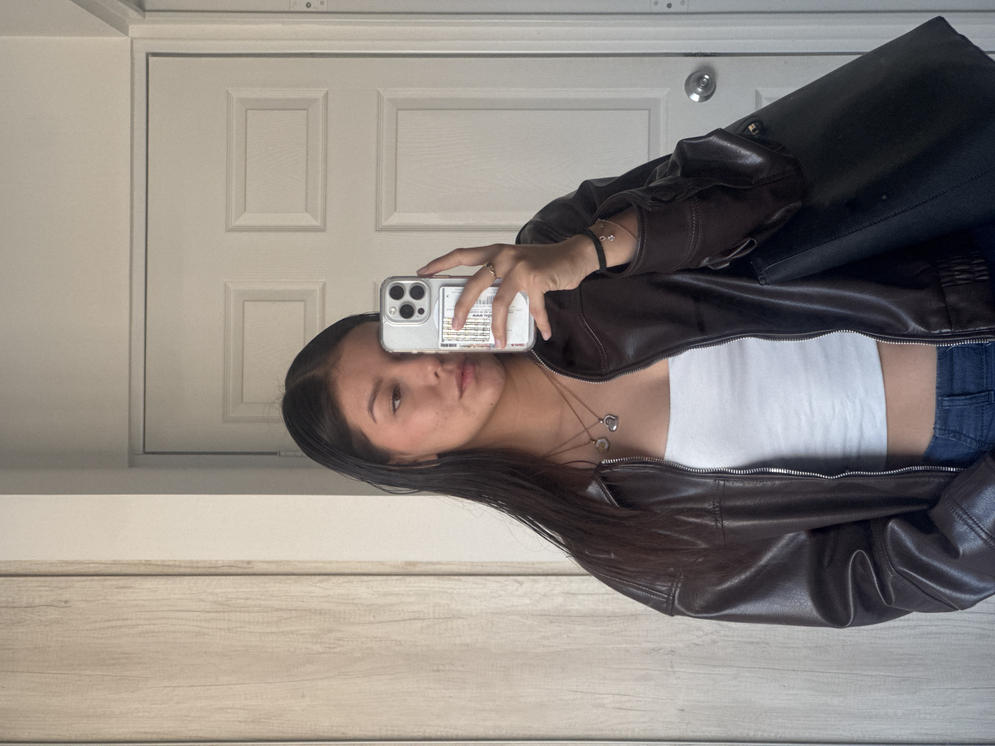 A young woman is captured in a mirror selfie, holding her smartphone to take the picture. She has long, dark hair and is wearing a dark brown leather jacket over a white crop top, paired with blue jeans. Around her neck are two layered necklaces, one featuring a small heart charm and the other a round pendant. She also wears a bracelet on her right wrist and a ring on her right hand.
The setting appears to be indoors, with a white paneled door and a silver doorknob visible in the reflection behind her. A light wood-textured floor or cabinet is also reflected below her. She has a neutral expression while taking the photo. A black bag or backpack is slung over her left shoulder, also visible in the mirror.
The smartphone she is using has a clear case, and a card with some printed text, including "www." and other alphanumeric characters, is visible behind the phone in the case. The image appears to be taken during the daytime due to the bright lighting. This media file is contextualized as potentially being from San Francisco Ocotlán (Ocotlán), Mexico.