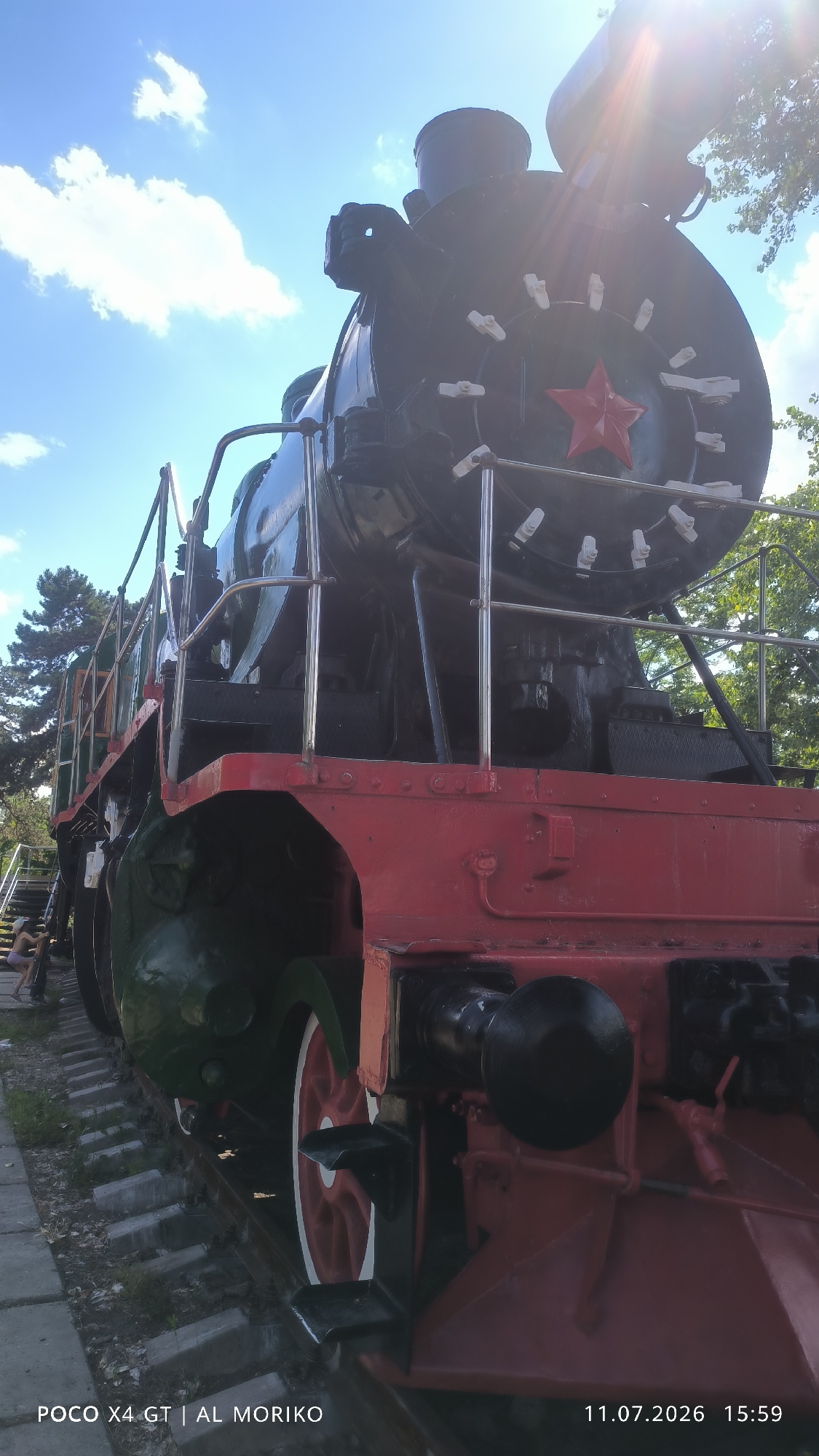 This is a low-angle, ground-level shot of the front of a vintage red and black steam locomotive. The locomotive is positioned on railway tracks in a sunny outdoor environment. The sky above is blue with scattered white clouds, and sunlight streams in from the upper right, creating lens flare. The locomotive features a prominent red star emblem on its front, surrounded by white-capped bolts. The engine has large wheels, one of which is visible with red and white spokes. The scene appears to be a public space, possibly a park or museum, as indicated by the presence of a paved walkway and some greenery. The bottom of the image displays text indicating the device used to capture the photo, "POCO X4 GT | AL MORIKO," and the date and time, "11.07.2026 15:59." The text suggests the location is Bender, Moldova.