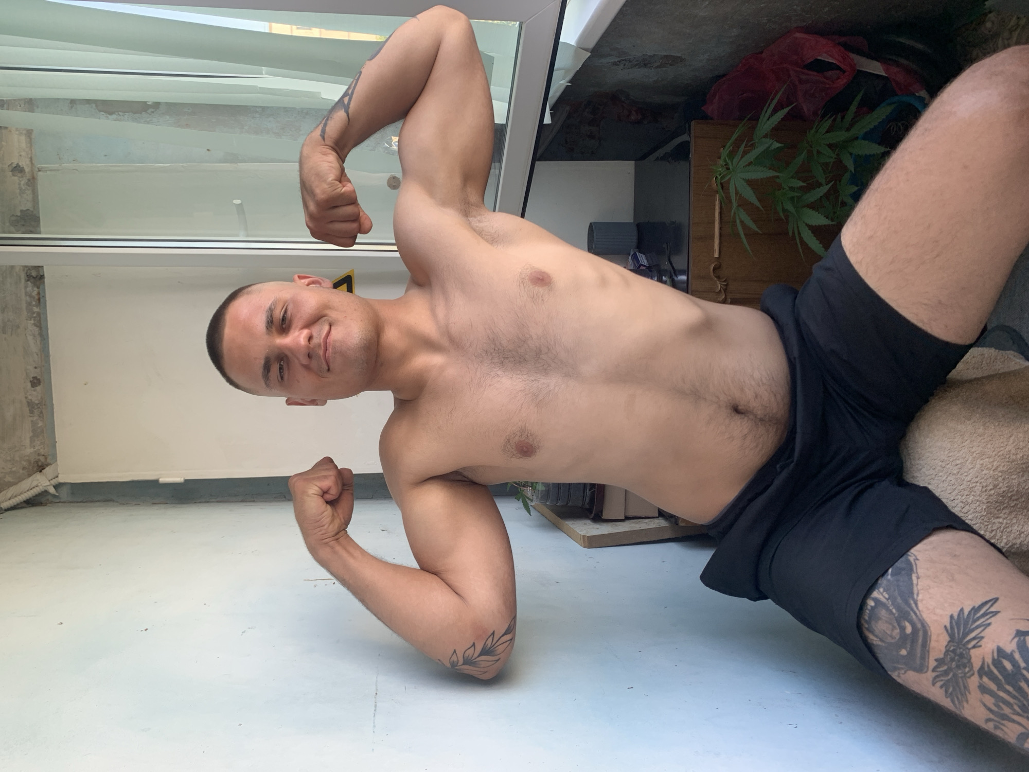 A shirtless man with a buzz cut, wearing black shorts, poses for a photo while flexing his biceps. He is seated on a chair or couch in what appears to be a balcony or an outdoor enclosed space with a light-colored wall on the left and a glass door with curtains on the right. In the background, there is a wooden cabinet with a potted plant, possibly cannabis, and some bags. The man has tattoos on his arms and legs. The lighting suggests it is daytime. The image does not provide any specific information about the location in the city of Pivdennoukrainsk, Ukraine.