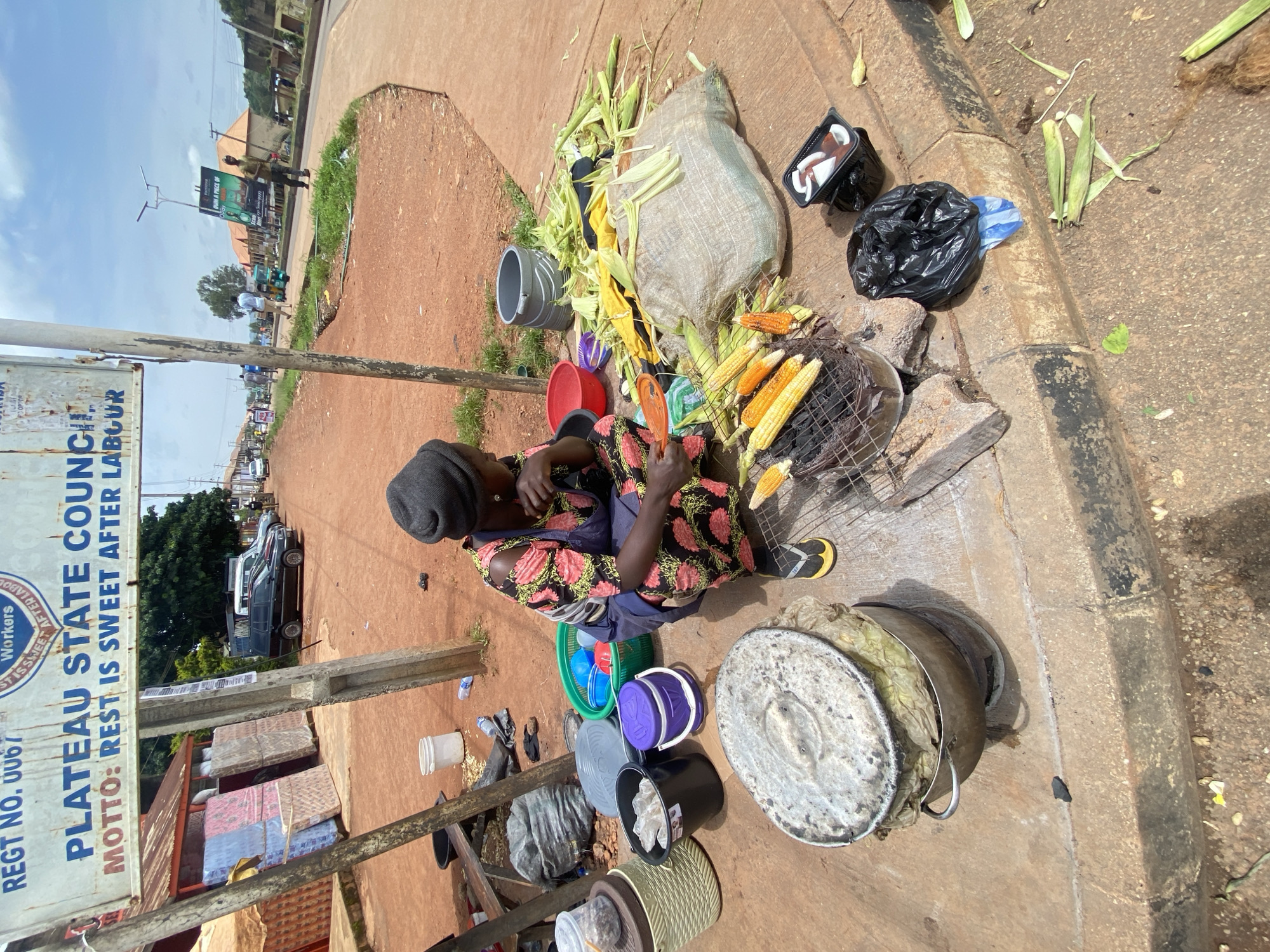 A woman wearing a black head covering and a colorful floral dress is crouching on a sidewalk in Jos, Nigeria, attending to a charcoal grill laden with corn. She appears to be preparing the corn for sale, with several ears already roasted and others waiting their turn. Surrounding her are various items related to her business, including husked corn, a large sack, buckets, and cooking utensils. In the background, a sign overhead reads "PLATEAU STATE COUNCIL" and "MOTTO: REST IS SWEET AFTER LABOUR," indicating the location as a public area within the Plateau State. The scene suggests a typical street-side vendor operation in a bustling urban environment.