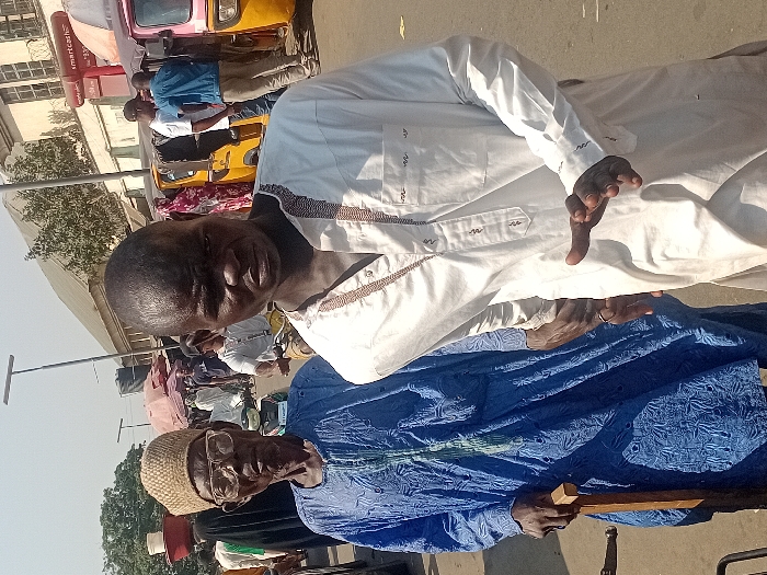 Two men pose for a photo in an urban environment. One man wears a bright white, long-sleeved garment, while the other is dressed in a blue traditional outfit, complete with a woven cap and eyeglasses. The older man holds a wooden staff. In the background, blurred images suggest a bustling street scene with vehicles and other people. The photo appears to be taken during the day.