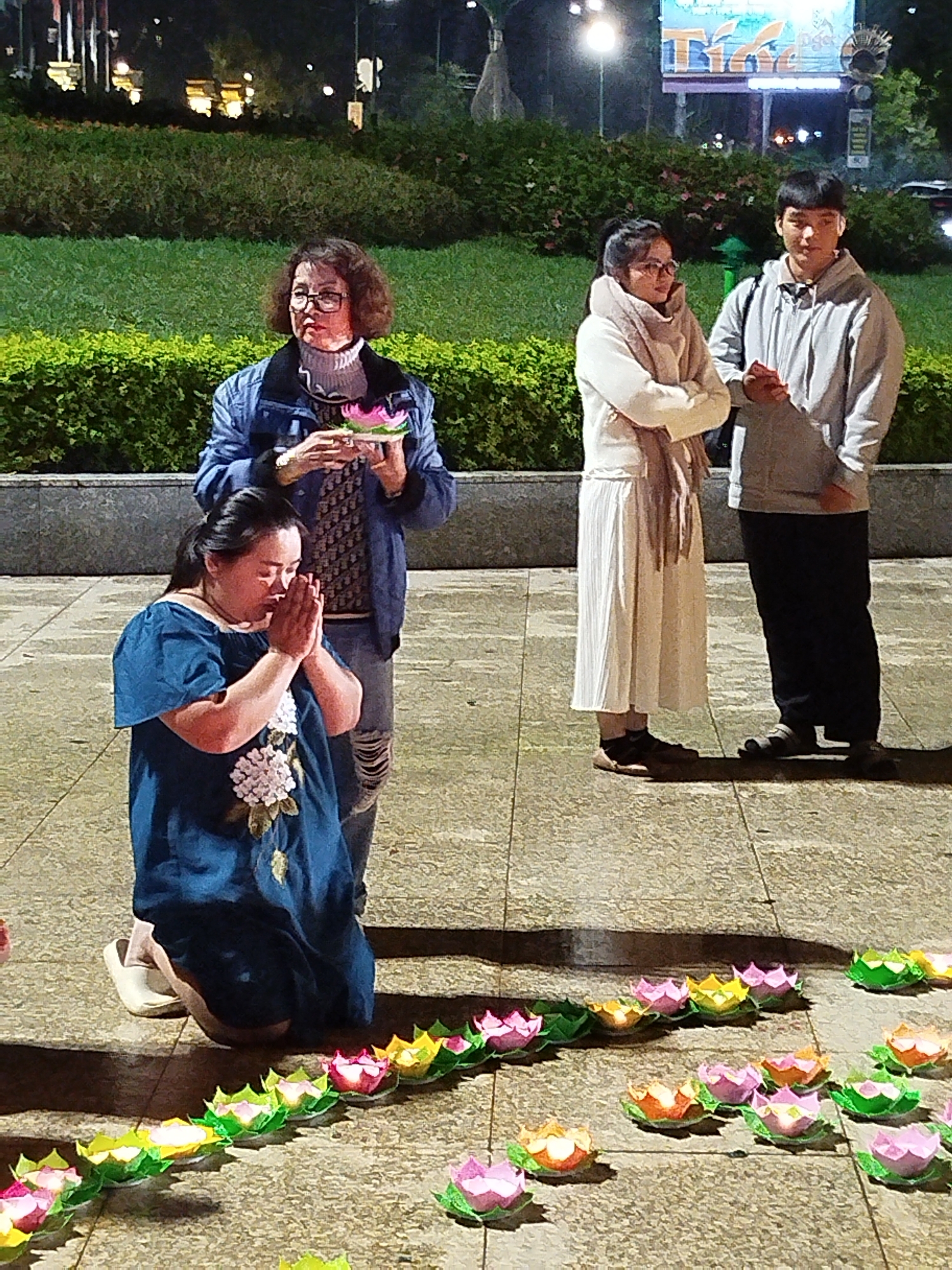 The image shows a woman kneeling in prayer among floating paper lotus lanterns on a stone surface, likely at night. She is wearing a blue dress with floral embroidery and white shoes. Behind her, an older woman in a denim jacket and jeans holds a lotus lantern. To the right, a young couple stands together, the woman in a white dress and scarf, and the man in a grey hoodie and black pants. In the background is a green hedge and a billboard. The location is Da Lat, Vietnam.