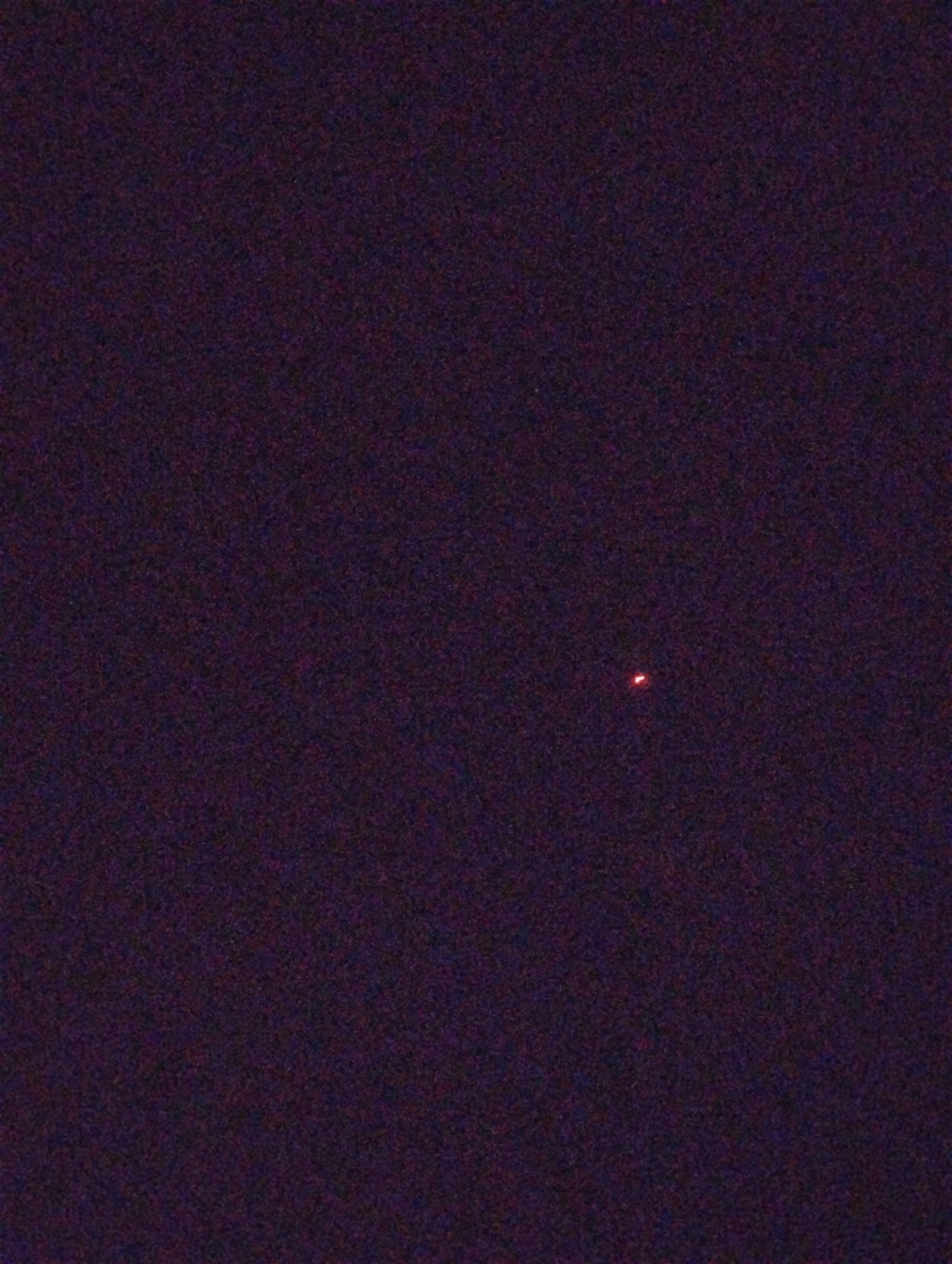 A small, bright red light source is visible against a dark, speckled background that appears to be the night sky. The light source is slightly elongated horizontally and is the only discernible feature in the image. The background is uniformly dark with a reddish-purple hue, indicative of low light conditions and potentially digital noise or atmospheric interference.