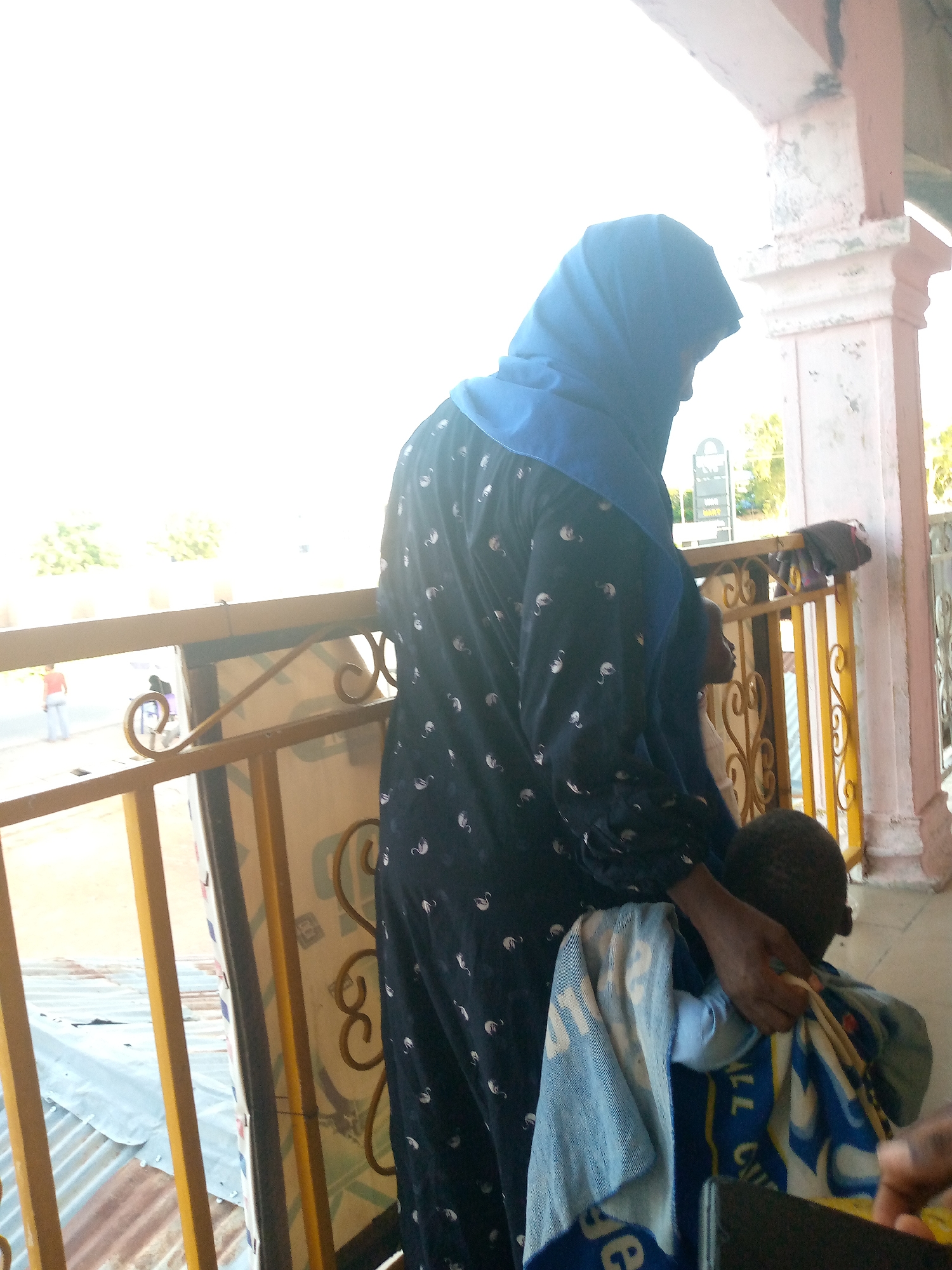 A person wearing a blue hijab and a dark, patterned dress stands facing away from the camera, holding a child in their arms who is wrapped in a blue and white towel. They are standing on a balcony or porch with a metal railing. In the background, there are trees, a street with some people, and buildings. The scene appears to be in an outdoor setting in a city, possibly Jalingo, Nigeria, given the architecture and vegetation.