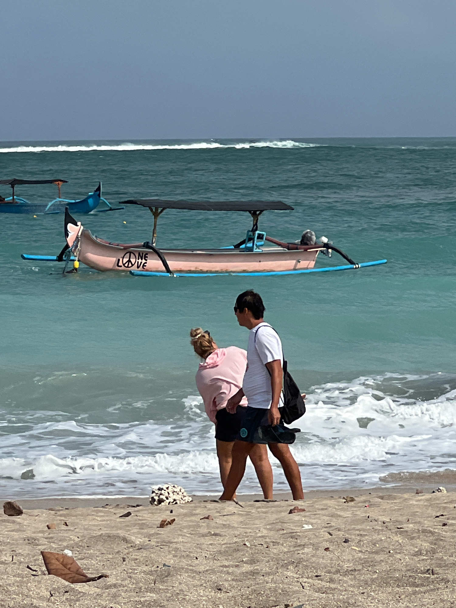 A medium shot captures a couple walking on the beach in Kuta, Bali, Indonesia. The woman, with blonde hair tied up, wears a pink hooded sweatshirt and black shorts. The man, with dark hair, is dressed in a white t-shirt and dark shorts, carrying a black backpack over his shoulder and a pair of dark flip-flops in his hand. They are walking away from the camera towards the water.
In the background, the turquoise ocean waves are breaking gently on the shore. Two traditional Indonesian fishing boats are anchored in the shallow water. The boat closer to the foreground is pink and has a black canopy. A peace symbol and the word "LOVE" are visible on its side. A second blue boat is further out in the water. The sky is overcast and a hazy blue. The beach is sandy with scattered debris, including shells and leaves.
