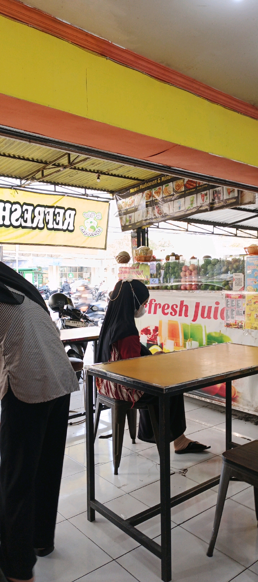 The image shows an outdoor food stall in Sidoarum, Indonesia. The stall is decorated with yellow and orange banners and has a corrugated metal roof. In the foreground, a person wearing a black hijab and a face mask sits at a table, facing away from the camera. A second person, also wearing a hijab and a striped shirt, stands to the left, with their back to the camera. Behind them, there is a display of fresh fruits and juices, with signs advertising "Menu Makanan & Minuman" (Food & Drink Menu) and "fresh Juic." The stall appears to be located along a busy street, with motorcycles and other vehicles visible in the background. The overall scene depicts a typical Indonesian street food establishment.