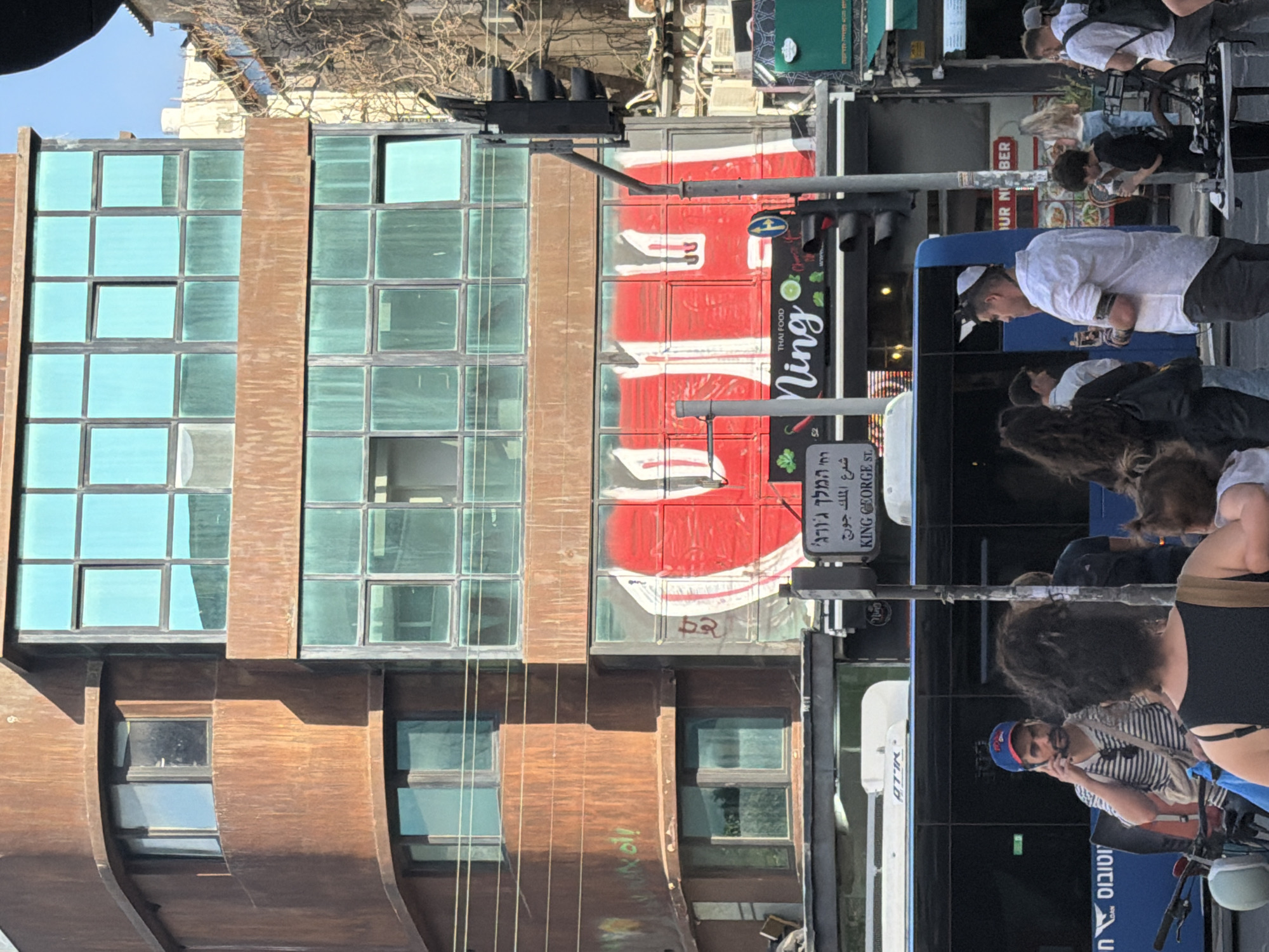 This image captures a vibrant urban street scene in Tel-Aviv, Israel, under bright, clear daylight. The main subjects include people, buildings, and a bus, indicating typical city activity.
To the left, a multi-story building features a facade of rusty brown, possibly corten steel, panels with numerous square windows, some tinted turquoise. Adjacent to it, another building presents a striking red and white surface with a large, partially obscured red circular element, possibly a logo or graffiti, and the number "52." Traffic lights on an overhead arm are visible against the sky.
At street level, several individuals are present. A man wearing a white t-shirt and a white baseball cap stands near a blue bus, looking to his left. Another man, wearing a striped shirt and a blue helmet, is seen on a bicycle, appearing to be using a phone. Other pedestrians are partially visible in the background, including a person with curly hair. A large blue and white bus, possibly a public transport vehicle, occupies a significant portion of the lower right frame.
Notable details include various street signs. One prominent sign is trilingual, displaying "המלך ג'ורג'" (Hebrew), "شارع الملك جورج" (Arabic), and "KING GEORGE ST." (English), confirming the location context. Another sign advertises "THAI FOOD Ning." Partially visible text on the side of the bus includes "וטובוס" (Hebrew, part of "אוטובוס" meaning 'bus') and "ATIR". The overall scene suggests a bustling commercial area with ongoing pedestrian and vehicular traffic.