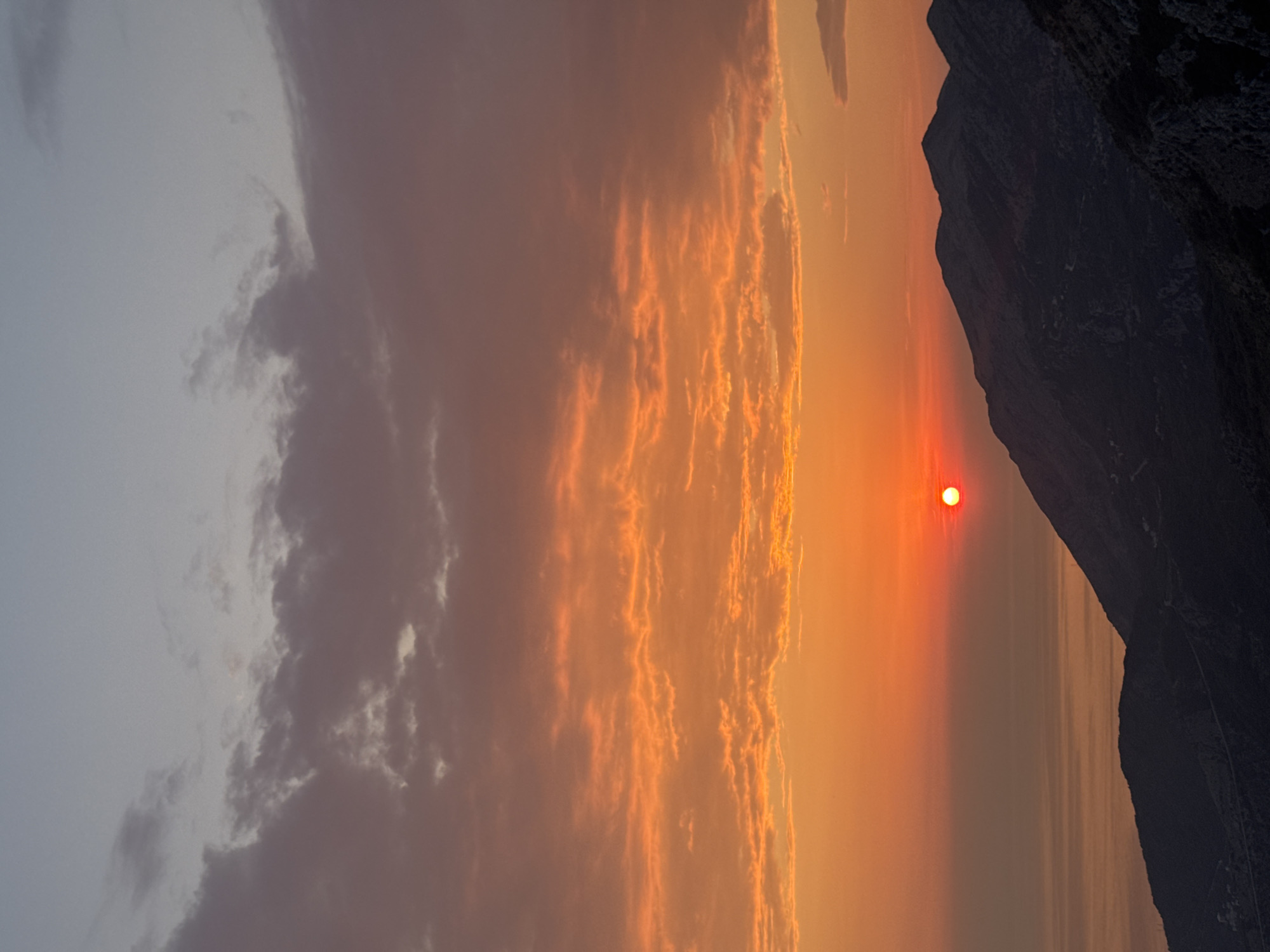 This is a vertical, medium-shot, outdoor photograph taken at sunset in Gluhi Do, Montenegro. The sun, a bright red orb, is low in the sky, casting a warm orange glow on the clouds and the sea. The sky transitions from a soft blue at the top to a vibrant orange near the horizon. Below the sky, dark, rugged mountains dominate the foreground, their slopes rendered in shades of deep blue and black. A winding road can be seen etched into the side of one of the mountains. The sea stretches out to the left, reflecting the colors of the sunset. There are no people or animals visible in the frame. The overall mood of the image is serene and majestic, capturing the natural beauty of the Montenegrin landscape at dusk.