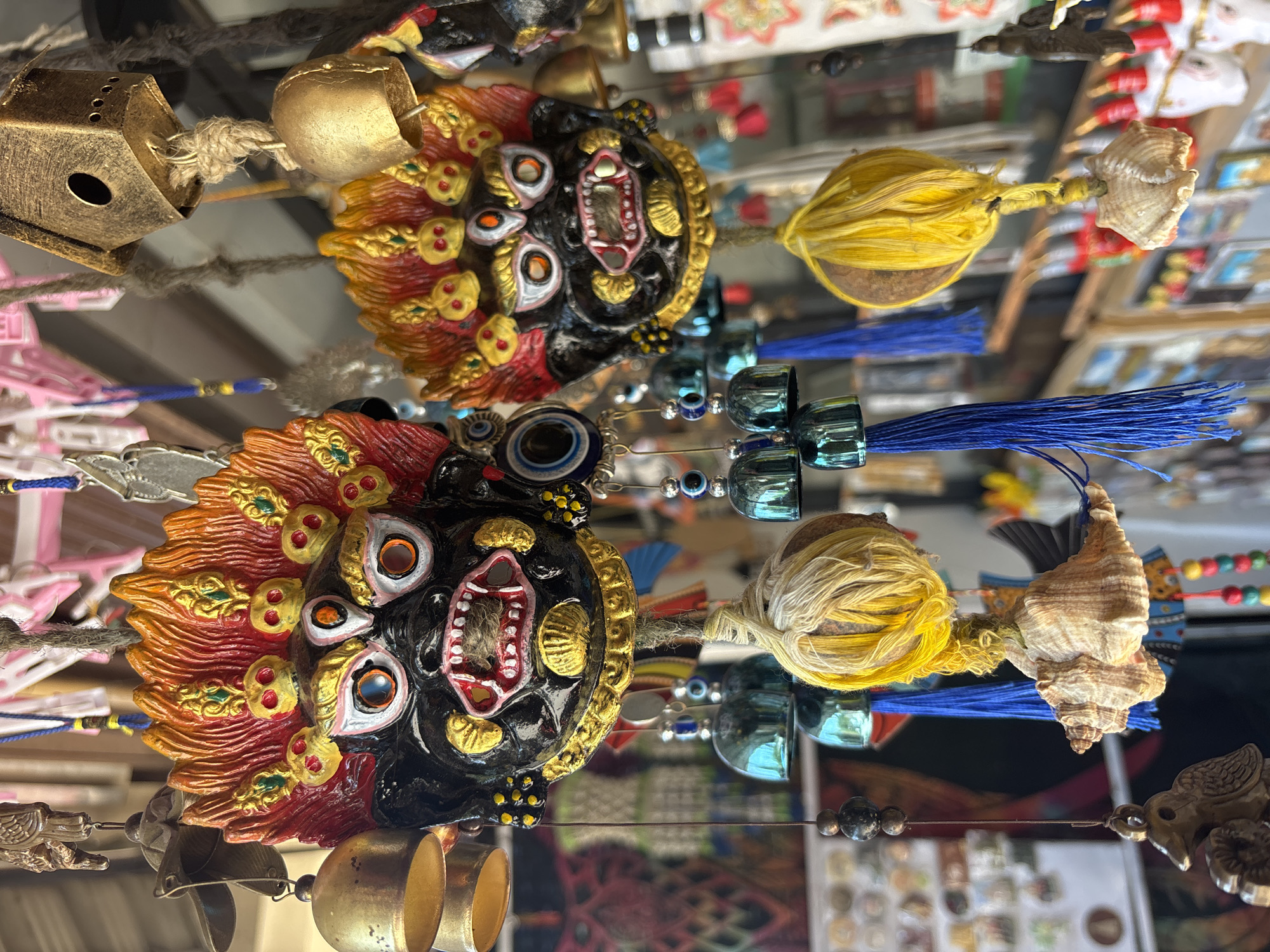 The image shows a reflection of a collection of decorative ornaments, possibly displayed for sale. The predominant ornament features a fierce mask with a dark face, a wide-open mouth with visible teeth, and multiple protruding eyes. A fringe of red and yellow “hair” surrounds the mask. Other elements include golden bells, blue beaded strings with tassels, a ball of yellow yarn, and a small seashell. The background suggests a shop or market setting with other items visible on shelves or display cases. Given the prompt's mention of Kamalapura, India, the scene likely depicts a shop selling local crafts or tourist souvenirs in that city.