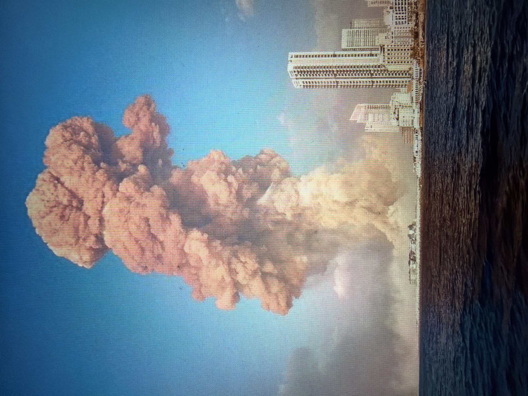 The image depicts a massive explosion, creating a towering mushroom cloud of dust and smoke that dominates the sky. The plume is a light brown and orange color, with a lighter, yellowish cloud emanating from its base. In the foreground, a dark blue body of water with moderate ripples occupies the bottom half of the image. Along the horizon, a city skyline is visible. The skyline features several tall, modern buildings, including at least one very tall skyscraper with many floors and windows. Other lower-rise buildings are also present. The explosion appears to have originated from the city's waterfront. The sky is a clear, bright blue.