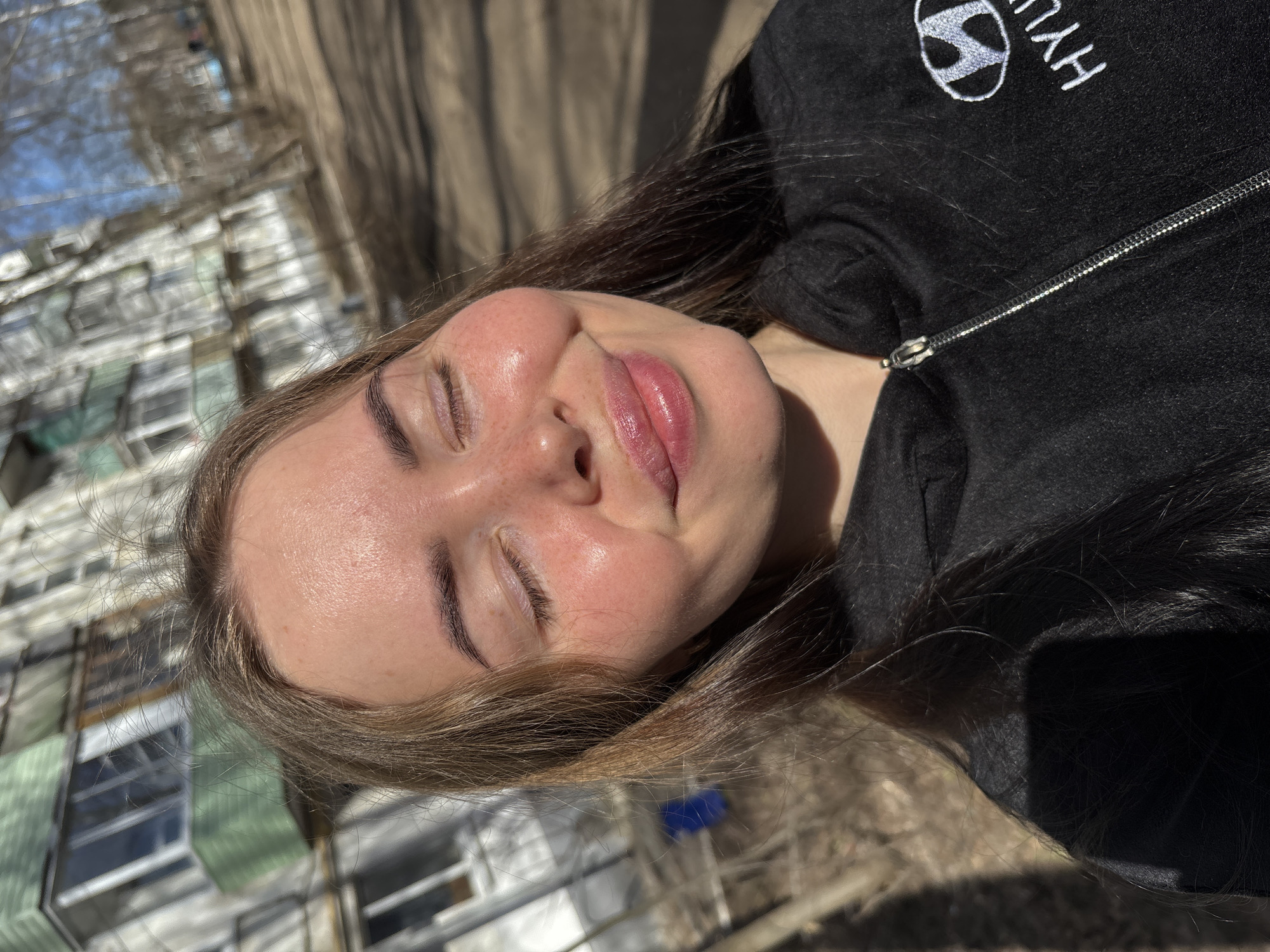 A young woman with light skin and closed eyes is lying outdoors in Izhevsk, Russia. Her brown hair is partially obscuring her face and is illuminated by sunlight. She is wearing a black zip-up hoodie with a "Hyundai" logo embroidered on the left side. The background includes a blurred view of buildings with green and white facades, suggesting an urban environment. Bare tree branches are also visible against a pale blue sky. The ground appears to be paved or covered with dry vegetation.
