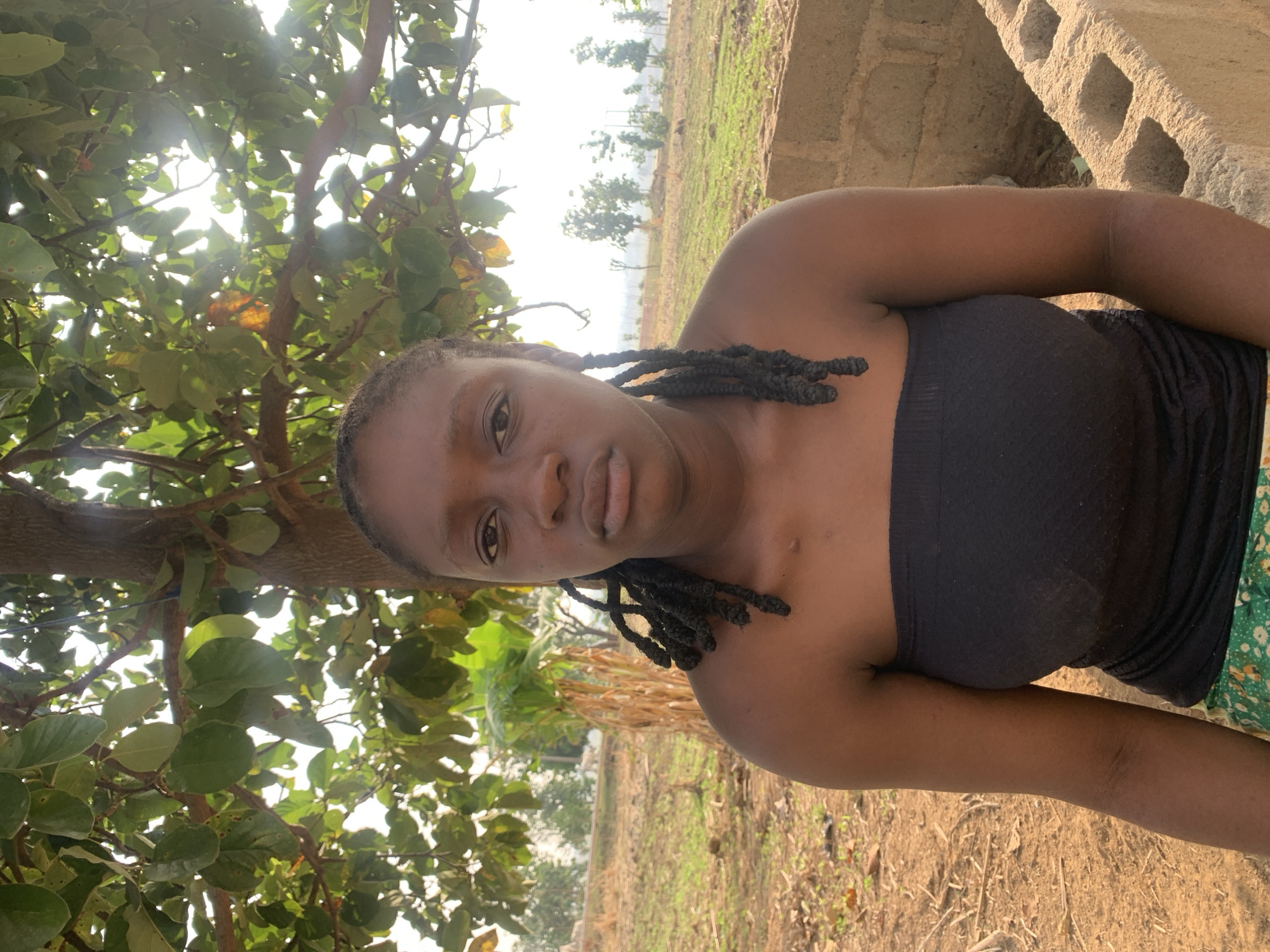A young woman with dark skin and braided hair looks directly at the camera, posing for a photograph. She is wearing a black strapless top, with her right arm visible at her side. Her expression is neutral and calm.
The setting is outdoors, likely in a rural or semi-rural area. Bright sunlight filters through the dense, green foliage of a tree, which dominates the left side and top of the frame. In the background, beyond the woman, there's a mix of dry earth and green fields, with some scattered trees in the distance. To the right, rough-hewn concrete blocks or a wall are visible. The overall scene suggests a sunny day with clear skies. No specific activity beyond posing is taking place, and no text is visible. This scene is set in Agban, Nigeria.