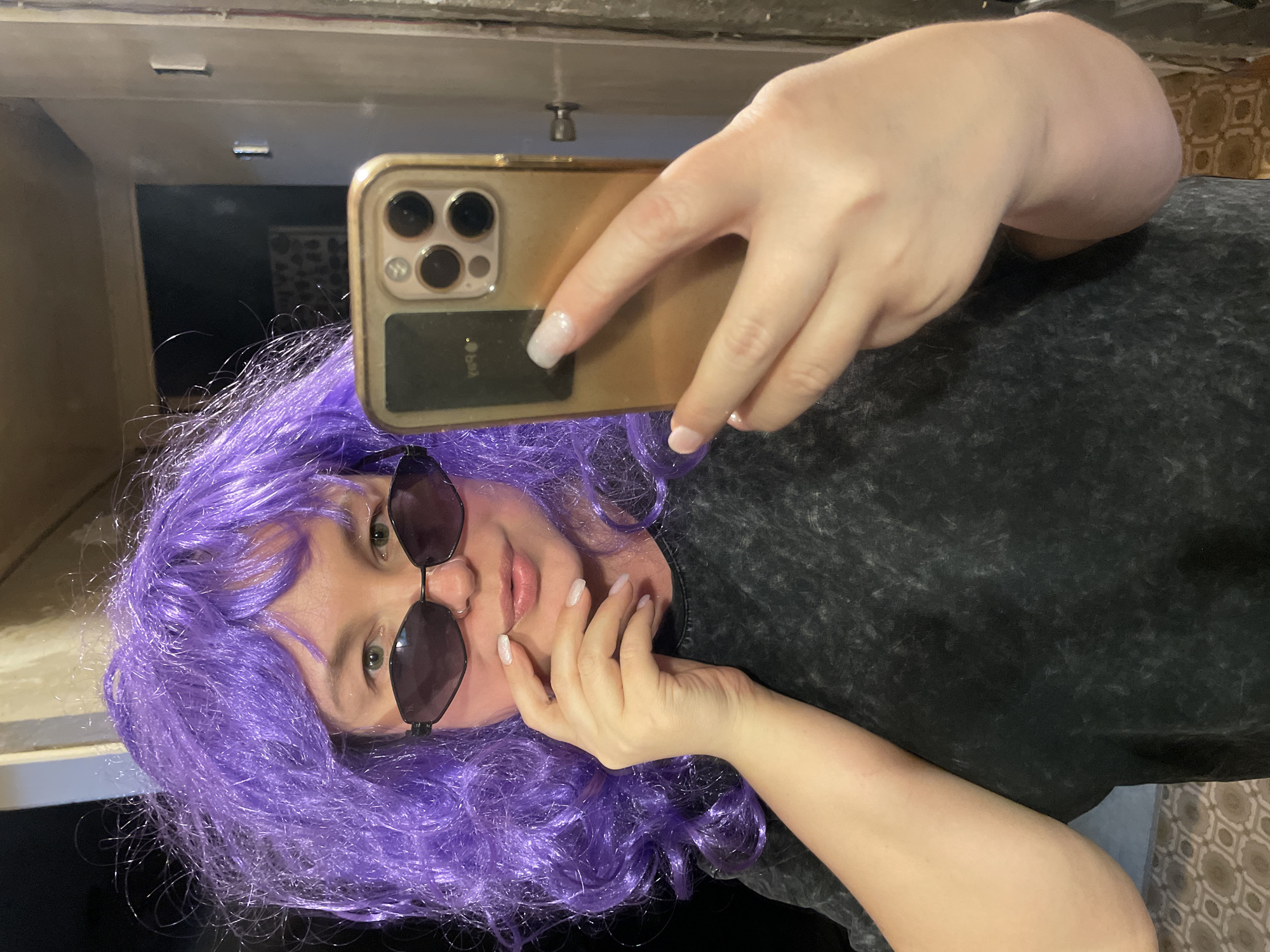 A woman with purple hair is taking a selfie with a gold-colored iPhone. She is wearing sunglasses and a black and gray shirt. She is standing indoors, possibly in a bathroom or hallway, as there is a mirror behind her. The specific location is not identifiable as Izhevsk, Russia, based solely on the visual information provided in the image.