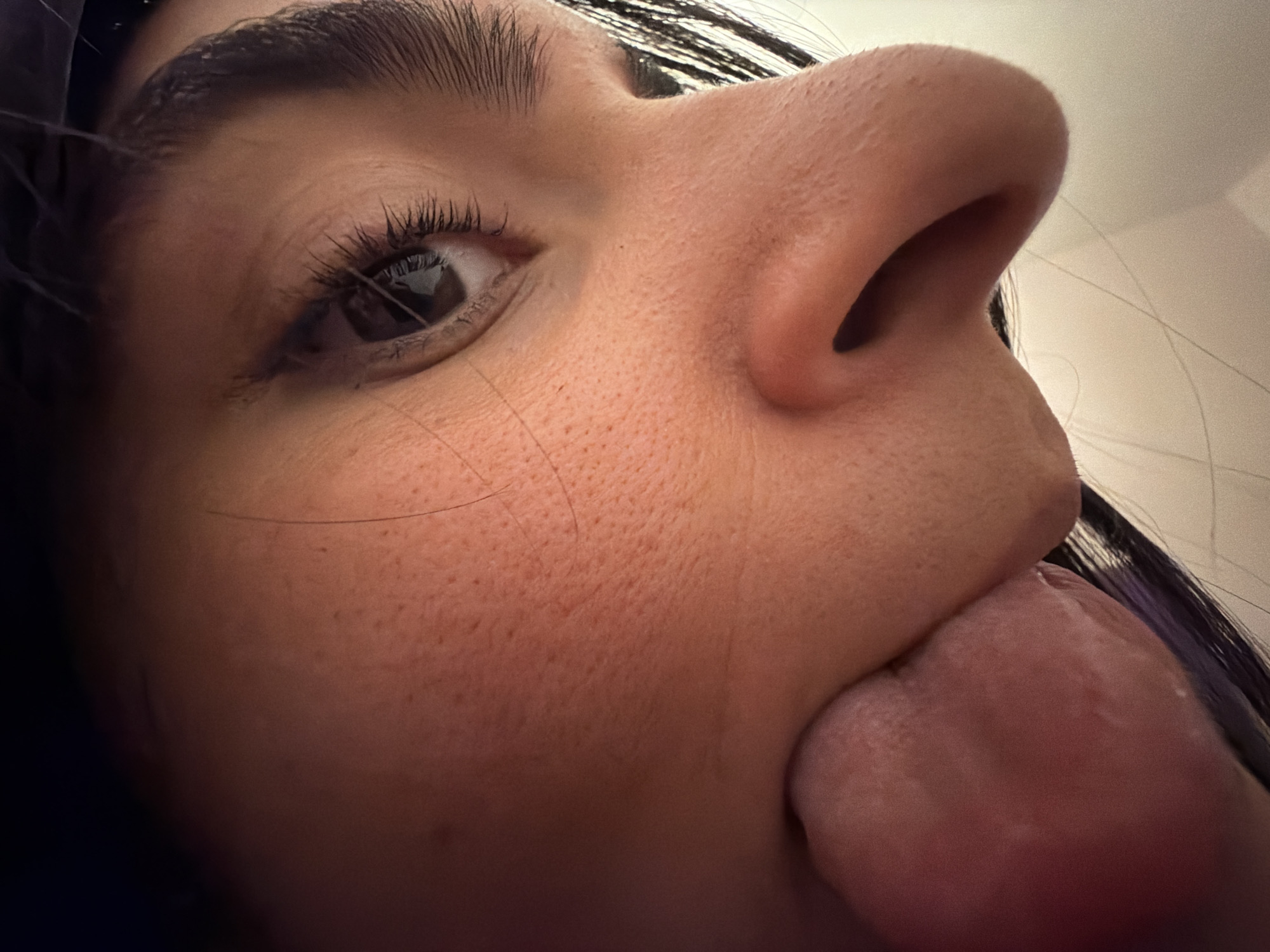 A close-up, distorted view of a person's face, taken from a very low angle. The subject's left eye is visible, with long eyelashes. The nose is prominent, filling a large portion of the frame. The person is sticking their tongue out. The background is blurred and appears to be a light color. There are strands of dark hair. There is no evidence in the image to indicate a location in New York City, United States of America.