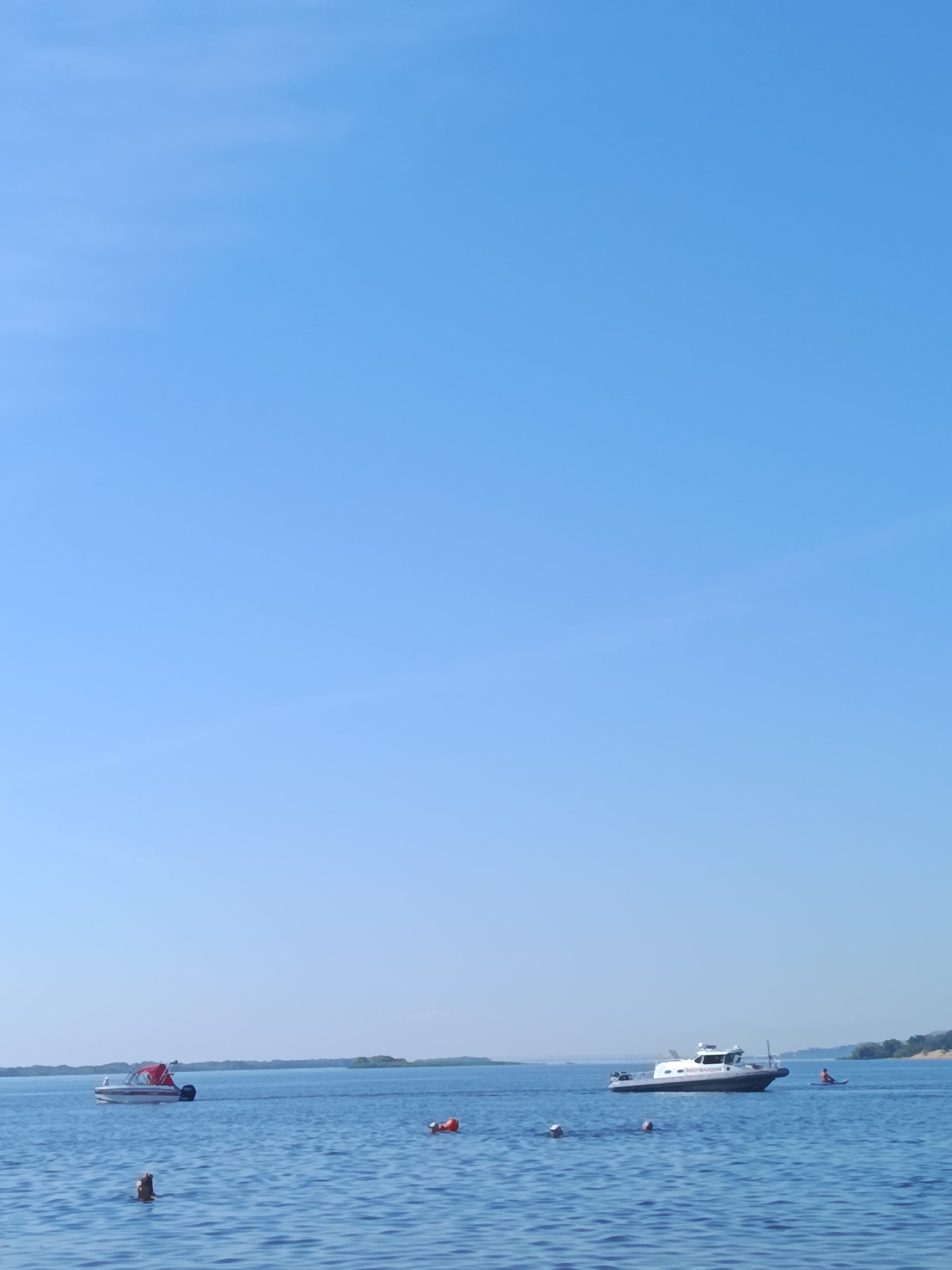 A wide shot captures a serene daytime scene on a body of water under a clear, pale blue sky. In the foreground, the calm, deep blue water is broken by the heads of several individuals swimming. A short distance away, an orange lifebuoy floats on the surface. To the left, a white boat with a red canopy is moving away from the viewer. Further out, a larger, white patrol boat with black accents is traveling across the water. To the right of the patrol boat, a person in an orange life jacket is visible, and further still, a person is on a small kayak. In the distance, a thin strip of land with scattered trees marks the horizon, suggesting a shoreline or a distant island. The overall impression is of a peaceful recreational activity taking place on a sunny day, likely in a location like the Volga River near Saratov, Russia, given the presence of patrol boats which are common for river safety.
