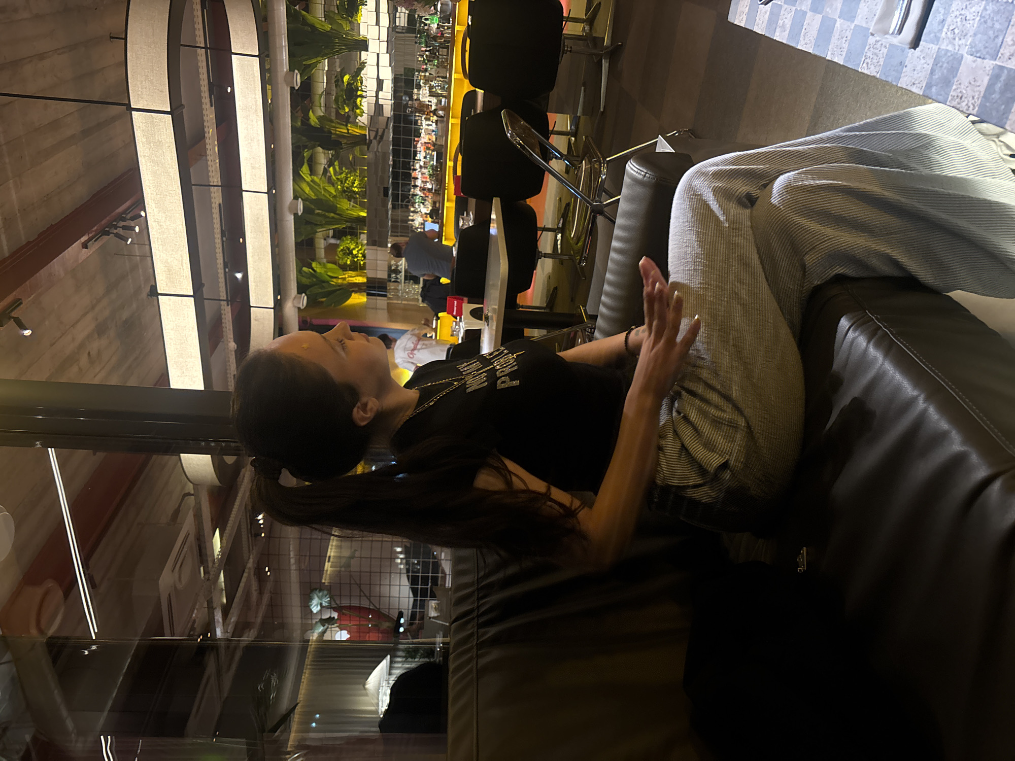 The image captures an indoor scene within a modern bar or restaurant, located in Yerevan, Armenia, as indicated by the context. A woman is prominently featured in the foreground, seated on a dark, leather-like couch. She is shown in profile, looking upwards and to the left, with her long dark hair pulled back in a ponytail. She wears a black short-sleeved top, on which the text "BALENCIAGA" is partially visible, and light-colored, striped trousers. Her hands are slightly raised, and her fingernails are painted a light color, suggesting she might be gesturing while speaking or lost in thought.
The background reveals a well-appointed bar area featuring a bright yellow counter. Black bar stools with metal frames are lined up along the counter. Behind the bar, shelves are stocked with numerous bottles and glasses, and abundant green plants add to the decor. Other indistinct figures of people can be seen in the background, including a person behind the bar.
The overhead lighting consists of large, arched fixtures with white fabric diffusers, casting a warm glow. Reflections of the interior are visible on a glass or reflective surface in the lower left, possibly a window or partition. A blue and white checkered floor pattern is also visible. The overall ambiance suggests an evening or nighttime setting, characterized by artificial lighting. The woman appears relaxed and engaged within this social environment.
