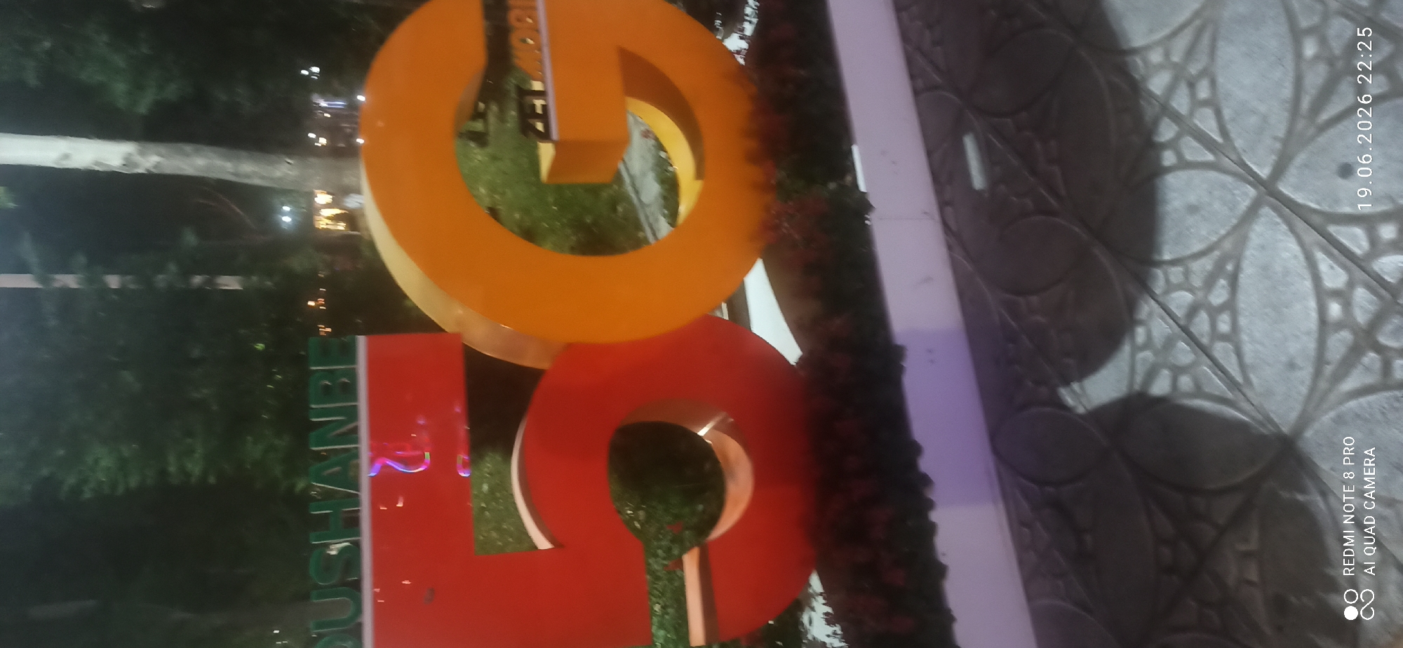 An outdoor, medium shot image taken at night depicts a large, red, three-dimensional number "5" and an orange, three-dimensional letter "G" in front of a blurry background of trees. Above and to the left of the "5" and "G", green lettering spells out "DUSHANBE". The red "5" is on the left, and the orange "G" is on the right. The objects are placed in front of a planter box filled with dark green foliage. The ground in the foreground has a patterned texture. The date "19.06.2026" and time "22:25" are visible in the bottom right corner, along with a "REDMI NOTE 8 PRO AI QUAD CAMERA" watermark.