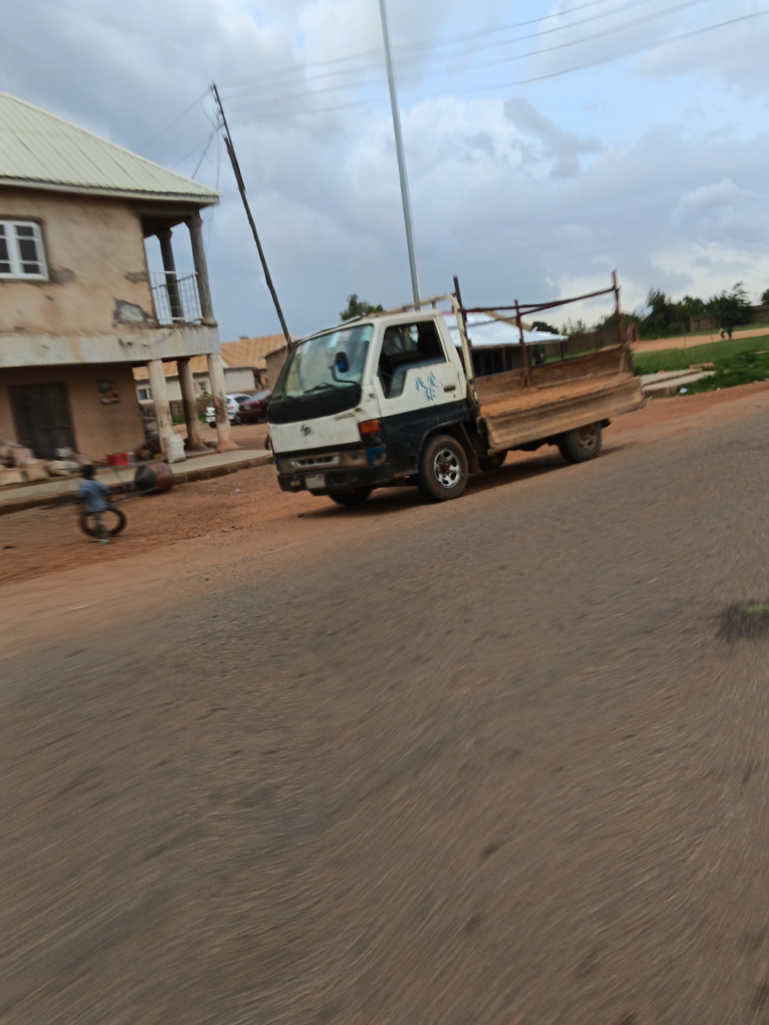 A light-colored truck with a wooden bed is parked on a dirt road next to a building with a light-colored roof. A person is standing and pushing a tire in front of the building. The sky is cloudy.