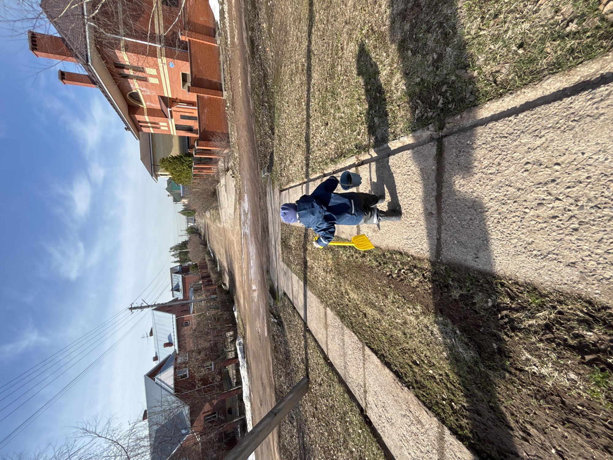 A small child in a blue snowsuit and hat plays on a concrete sidewalk next to a grassy area. The child has a yellow shovel and a blue bucket. The sidewalk runs alongside a dirt road, with houses visible in the background. Power lines are visible in the sky. The houses are primarily red brick, typical of suburban Russia. The location is possibly in the city of Ферма, Russia. The weather is sunny. The season appears to be early spring, as evidenced by the patchy grass and bare trees.